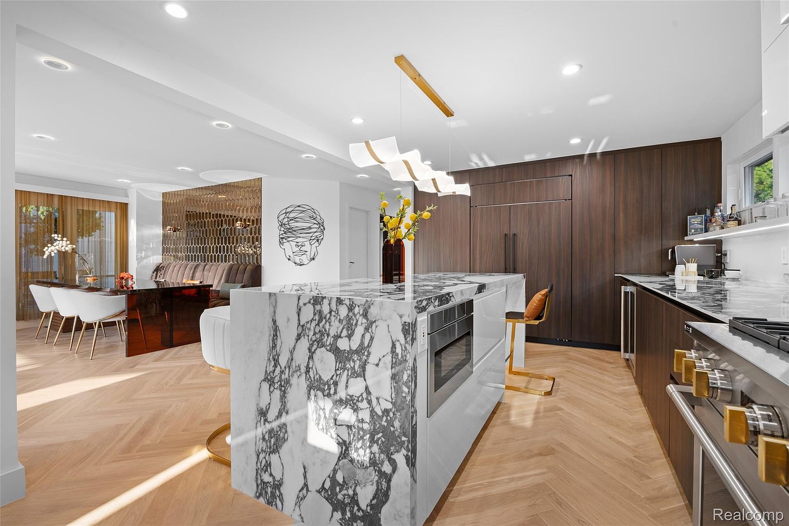 This is a modern kitchen featuring a large marble island with built-in appliances and seating. The kitchen also includes dark wood cabinetry, stainless steel appliances, and a dining area with a unique decorative screen. The herringbone wood flooring adds warmth and texture to the space.