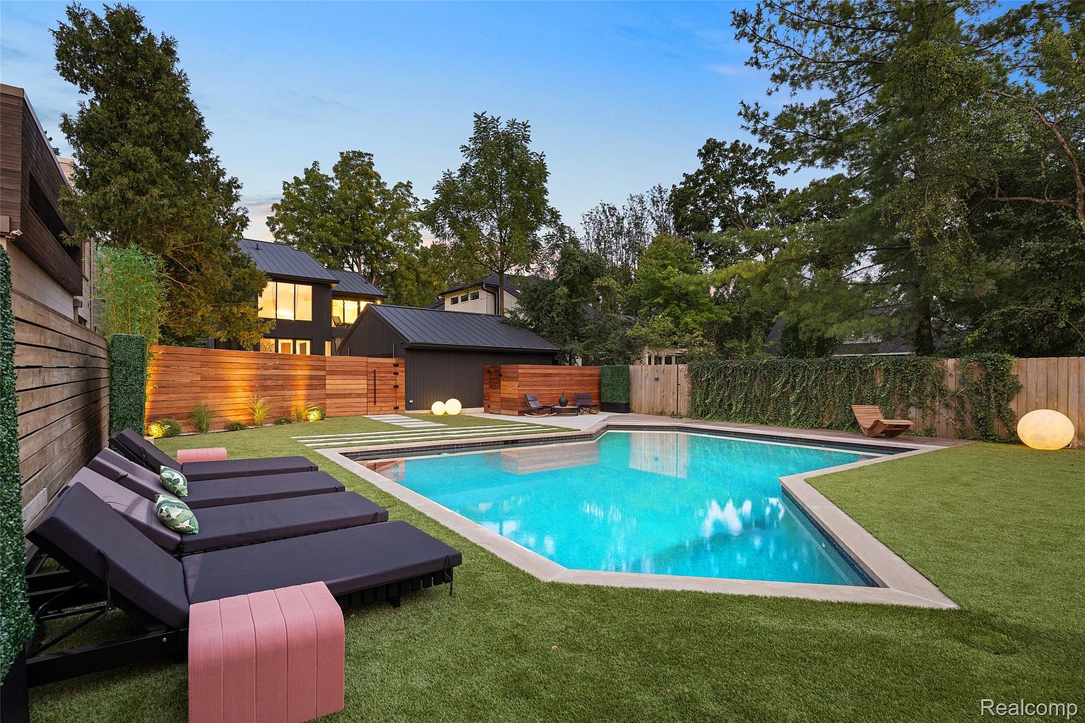 This image showcases a luxurious backyard pool area, featuring a modern geometric pool surrounded by well-manicured artificial turf. Several lounge chairs are arranged poolside, and the area is enclosed by a stylish wooden fence and lush greenery, providing privacy. The scene is set against a backdrop of a contemporary home with dark accents, creating an inviting and upscale outdoor living space.