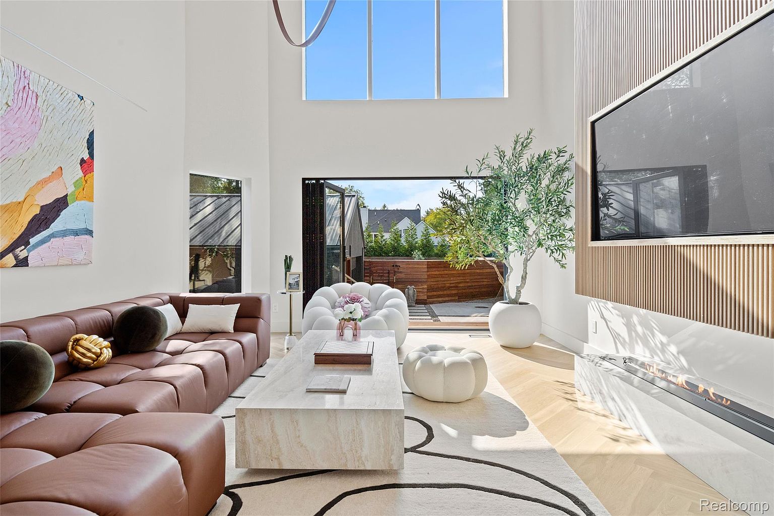 This is a bright and modern living room featuring a large brown sectional sofa, a light-colored stone coffee table, and a white area rug with black line design. The room has high ceilings with a large window above the folding glass doors that lead to an outdoor patio. A modern fireplace and a large television are mounted on a wood-paneled wall, and a potted olive tree adds a touch of nature to the space.