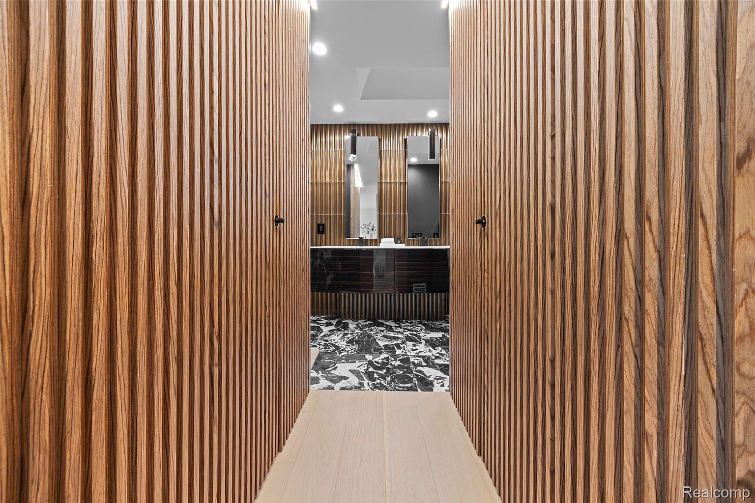 This image showcases a luxurious primary bathroom with a unique design. The entrance is framed by vertically paneled wooden doors, leading into a space with a striking black and white marble floor. The bathroom features a dark wood vanity with dual sinks and modern mirrors, complemented by textured wall panels and recessed lighting, creating a sophisticated and spa-like atmosphere.
