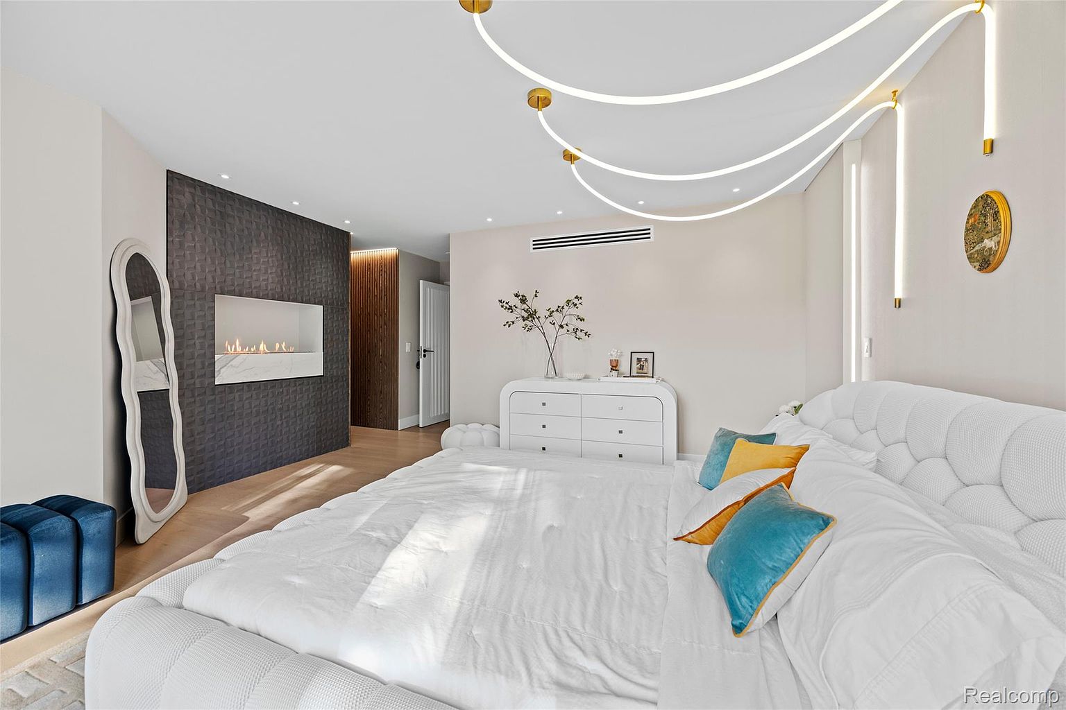 This is a primary bedroom featuring a modern design with a unique lighting fixture and a textured wall with a fireplace. The room includes a large bed with white linens and decorative pillows, a dresser, and a full-length mirror. The overall impression is luxurious and stylish, creating a relaxing and inviting atmosphere.
