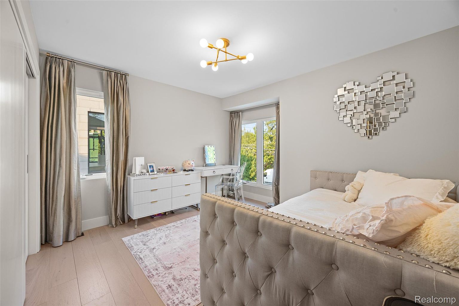 This is a well-lit bedroom featuring a neutral color palette with light wood flooring and soft gray walls. The room includes a tufted headboard bed, a white dresser with decorative items, and a vanity area with a clear chair. A heart-shaped mirror adds a decorative touch, and the overall impression is clean, stylish, and inviting.