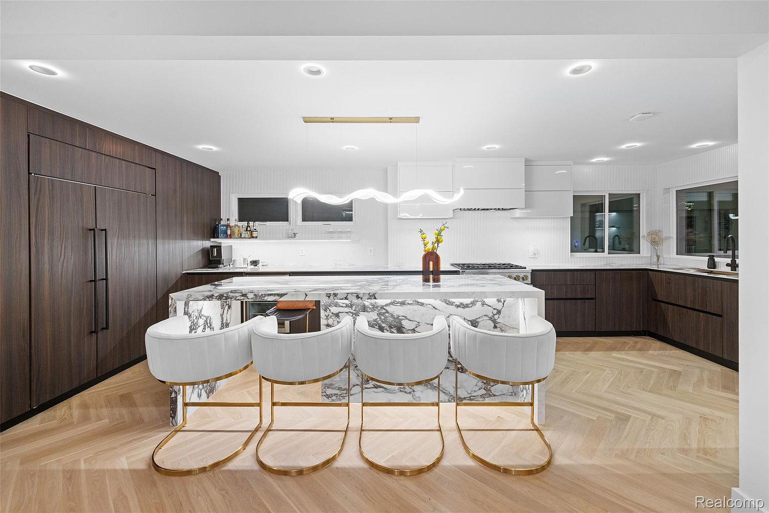 This is a modern kitchen with dark wood cabinetry and a large marble island with seating for four. The kitchen features white upper cabinets and a white backsplash, complemented by a unique modern light fixture above the island. The flooring is a light-colored herringbone pattern, adding warmth to the space.