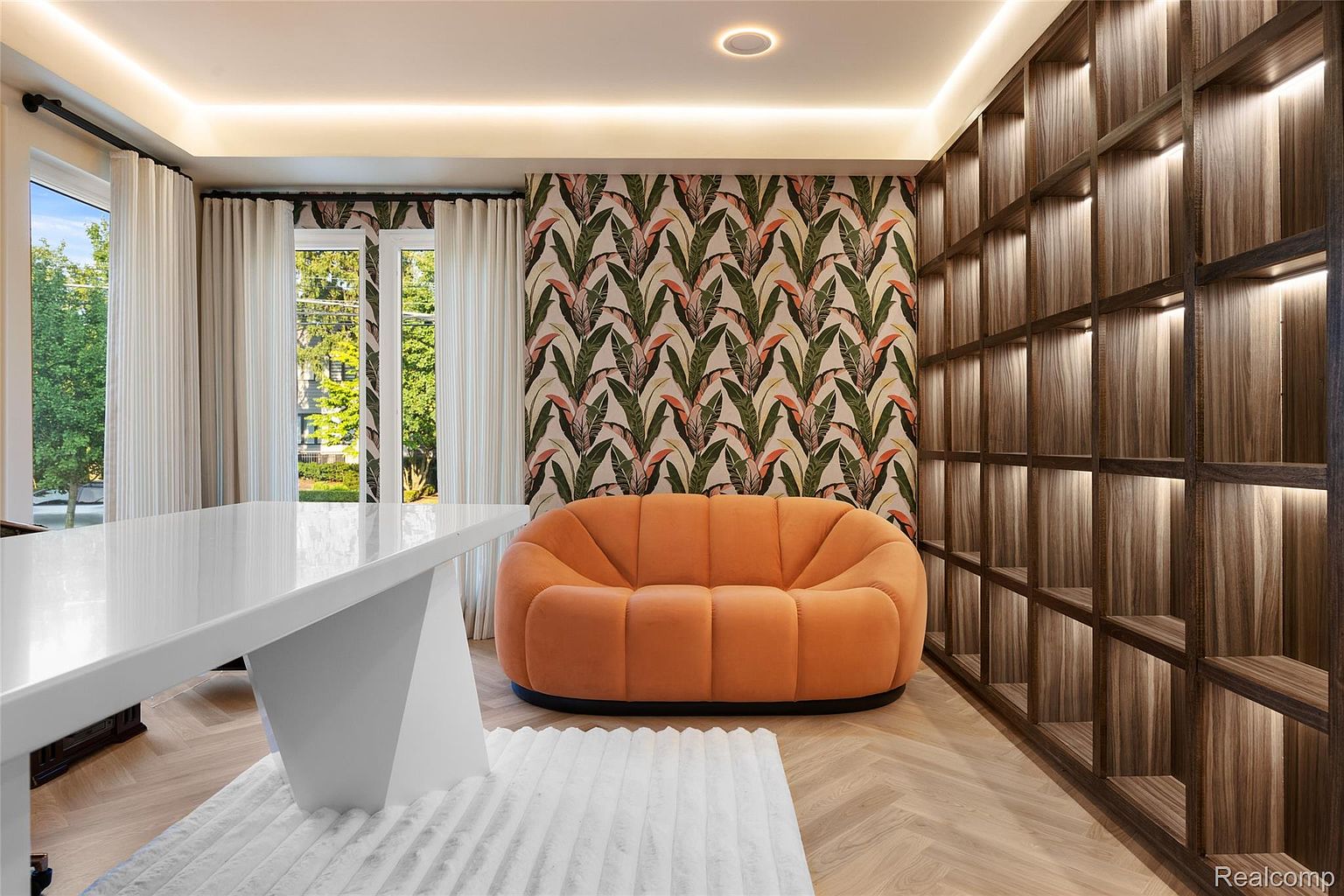 This interior shot showcases a stylish office or study area featuring a unique orange sofa against a backdrop of patterned wallpaper and a custom-built wooden shelving unit with integrated lighting. A modern white desk sits on a plush white rug, adding to the room's contemporary aesthetic. The herringbone wood flooring and natural light from the window create a warm and inviting atmosphere.