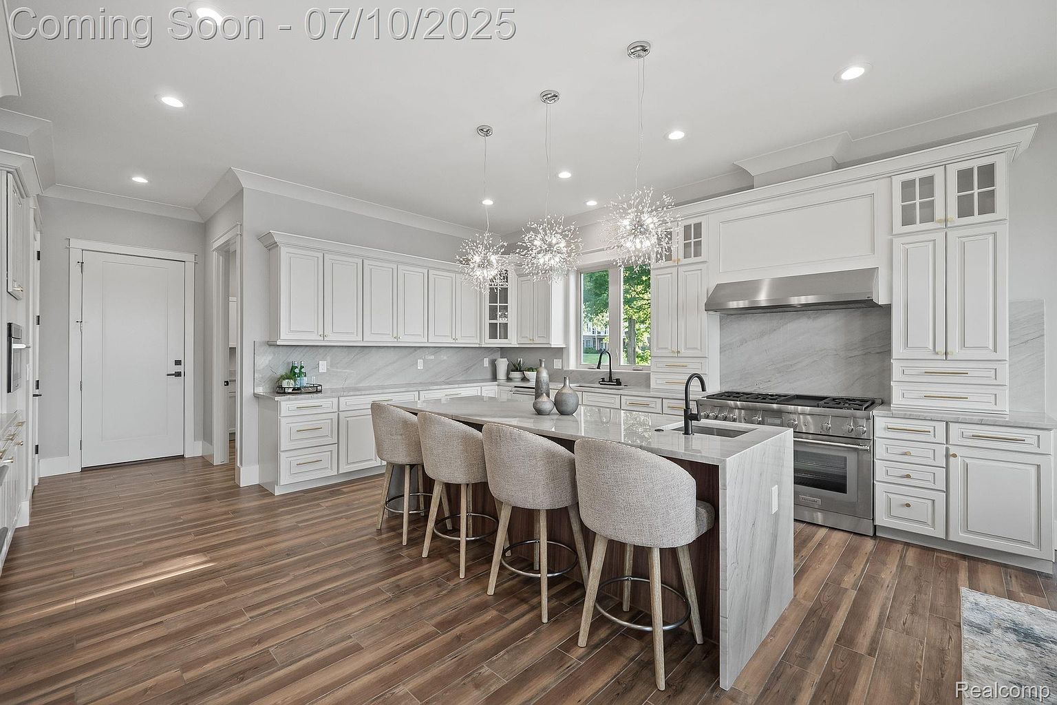 This is a bright and spacious kitchen featuring white cabinetry, marble countertops and backsplash, and stainless steel appliances. A large island with seating provides ample workspace and a focal point, while modern pendant lighting adds a touch of elegance. The hardwood flooring and natural light create a warm and inviting atmosphere.