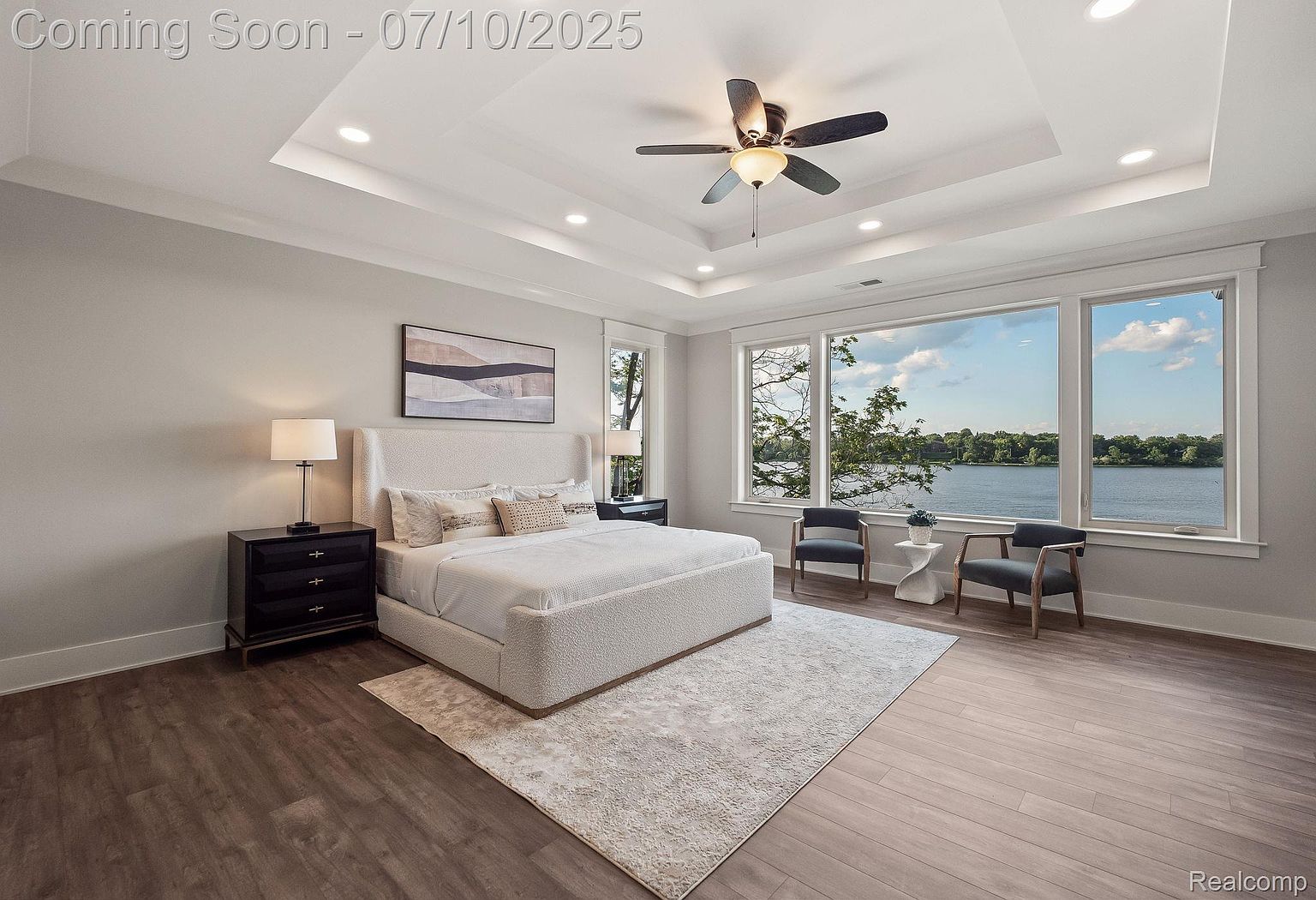 This is a well-lit primary bedroom featuring a large bed with a textured headboard and neutral bedding, complemented by dark wood nightstands and lamps. The room boasts a tray ceiling with recessed lighting and a ceiling fan, while a large window offers a scenic view of the water. The flooring is wood, and a light-colored area rug adds softness to the space.