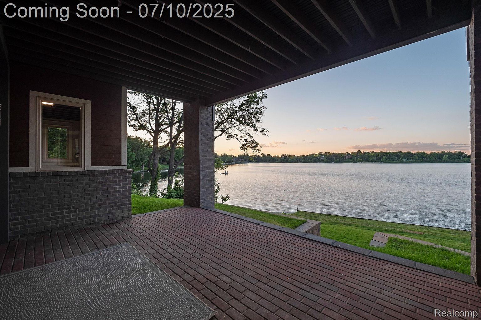 This image showcases a covered patio or balcony with a brick floor, offering a serene view of a lake. The structure features brick pillars and a dark-toned ceiling, creating a sheltered outdoor living space. The transition from the patio to the grassy lawn and the lake beyond enhances the property's appeal, suggesting a tranquil and picturesque setting.