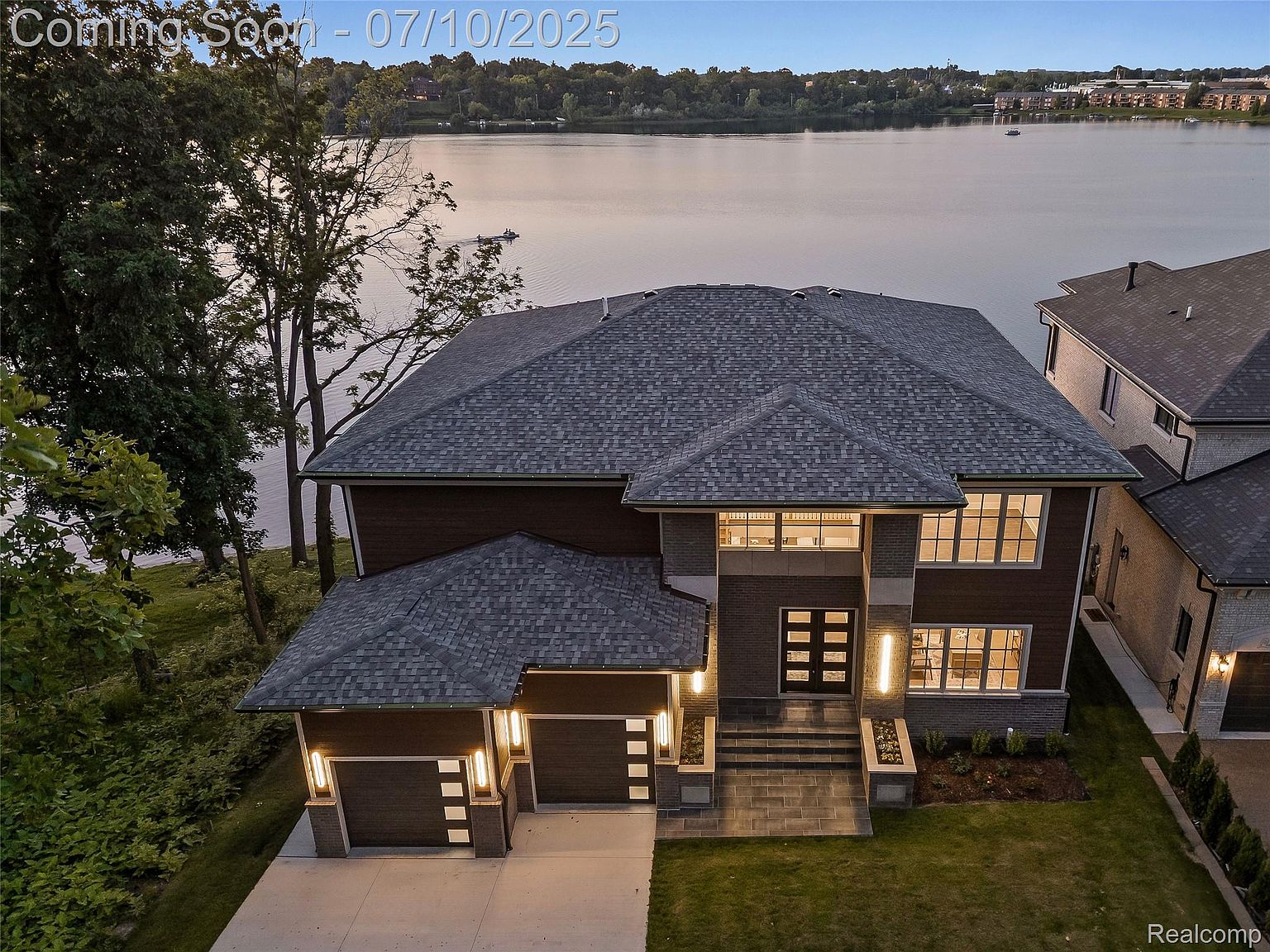 This is an aerial view of a luxurious two-story home situated on a waterfront property. The house features a dark gray roof, a brick facade, and a two-car garage. The well-manicured lawn and the serene lake in the background enhance the property's appeal, creating a sense of exclusivity and tranquility.