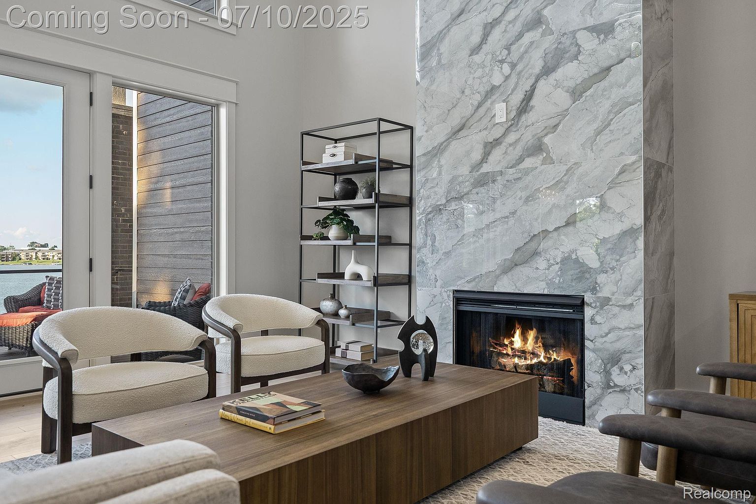 This living room features a modern fireplace with a marble surround, flanked by a dark metal shelving unit. Two cream-colored armchairs face a wooden coffee table, creating a cozy and inviting seating area. Large windows provide natural light and a glimpse of the outdoors, enhancing the room's appeal.