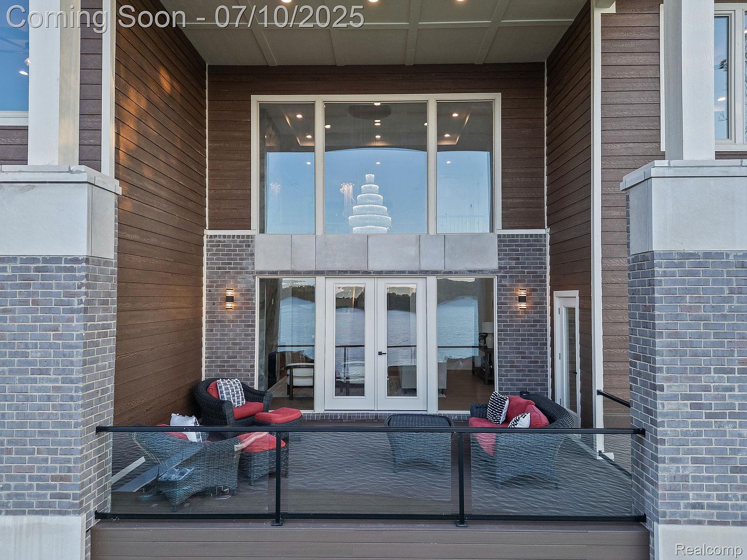 This image showcases a luxurious patio or deck area, featuring comfortable wicker seating with red cushions and a modern glass railing. The space is framed by the building's exterior, which includes a combination of brick and wood siding, along with large windows and doors that offer a glimpse of the interior and the view beyond. The overall impression is one of upscale outdoor living and relaxation.