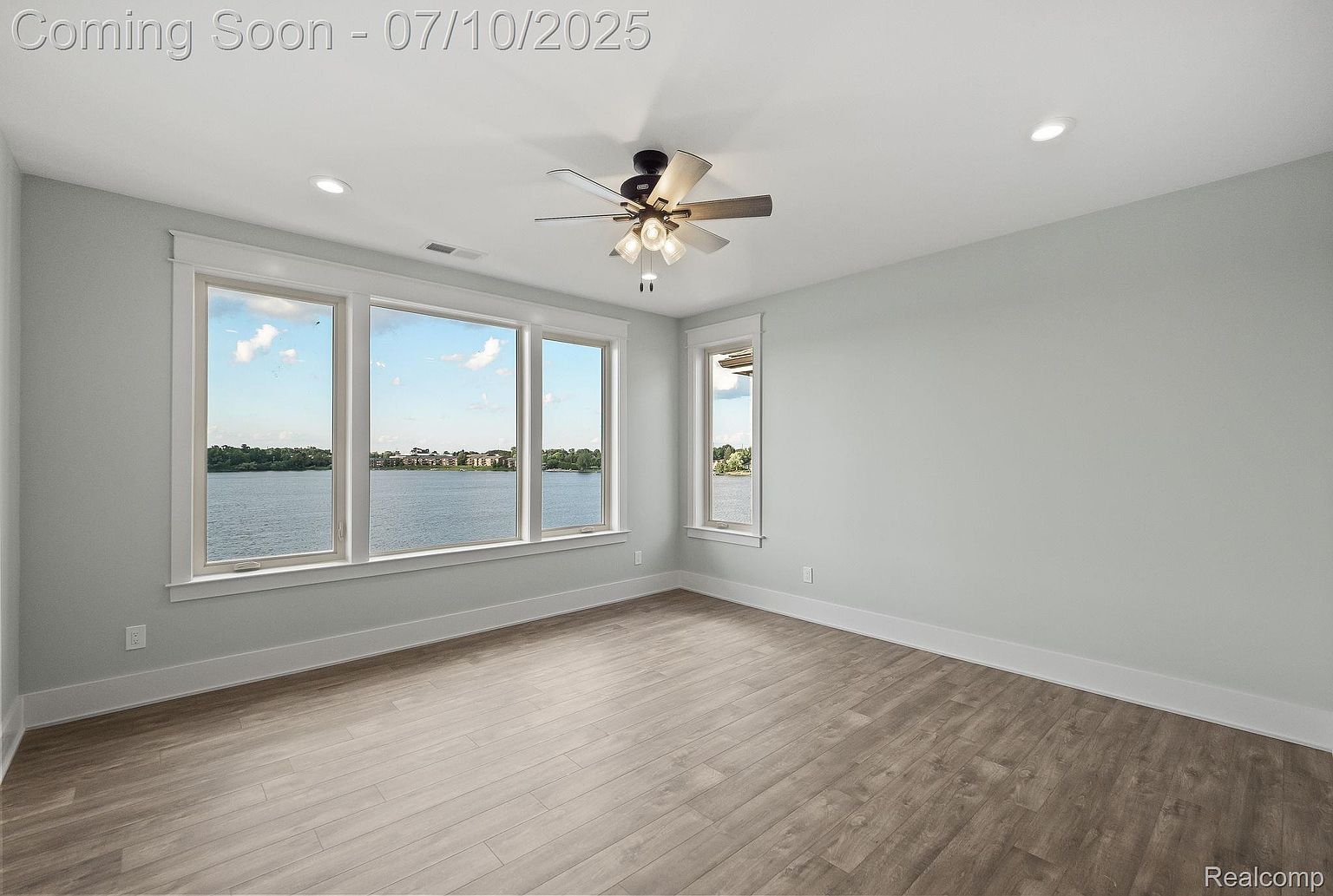 This is a bright and airy primary bedroom with a modern aesthetic. The room features large windows offering a scenic lake view, light gray walls, and wood-look flooring. A ceiling fan with integrated lighting adds a functional and stylish touch.