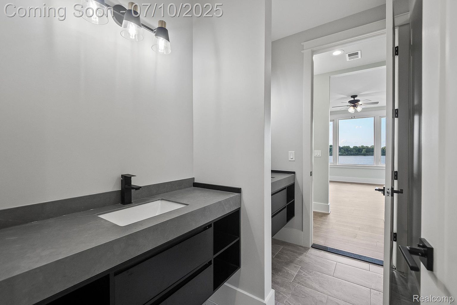 This is a modern primary bathroom featuring a sleek, dark vanity with a gray countertop and a contemporary faucet. The room has a minimalist design with clean lines and a neutral color palette, leading to an adjacent room with large windows overlooking a body of water. The flooring appears to be a light-colored tile, and the overall impression is one of sophistication and tranquility.