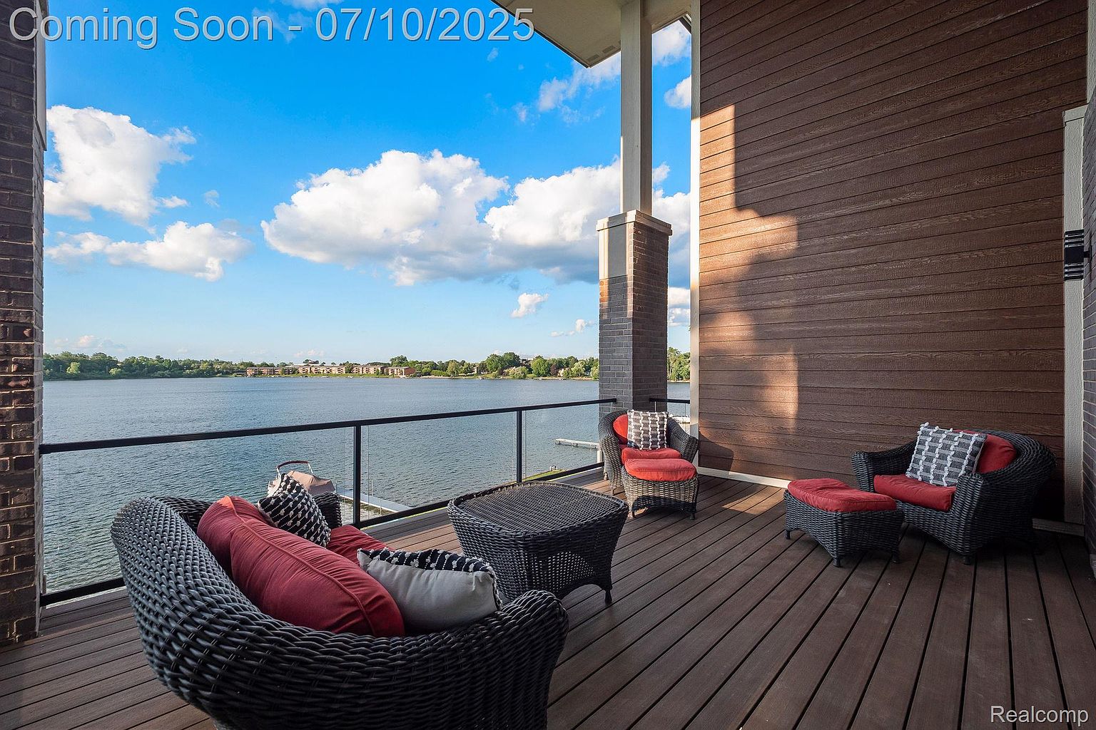 This is a beautiful outdoor patio/deck/balcony area with wicker furniture, including chairs, a table, and an ottoman, all arranged to create a comfortable seating area. The deck overlooks a serene lake, offering stunning water views and a sense of tranquility. The space is perfect for relaxation and entertaining, with a modern design and a focus on outdoor living.