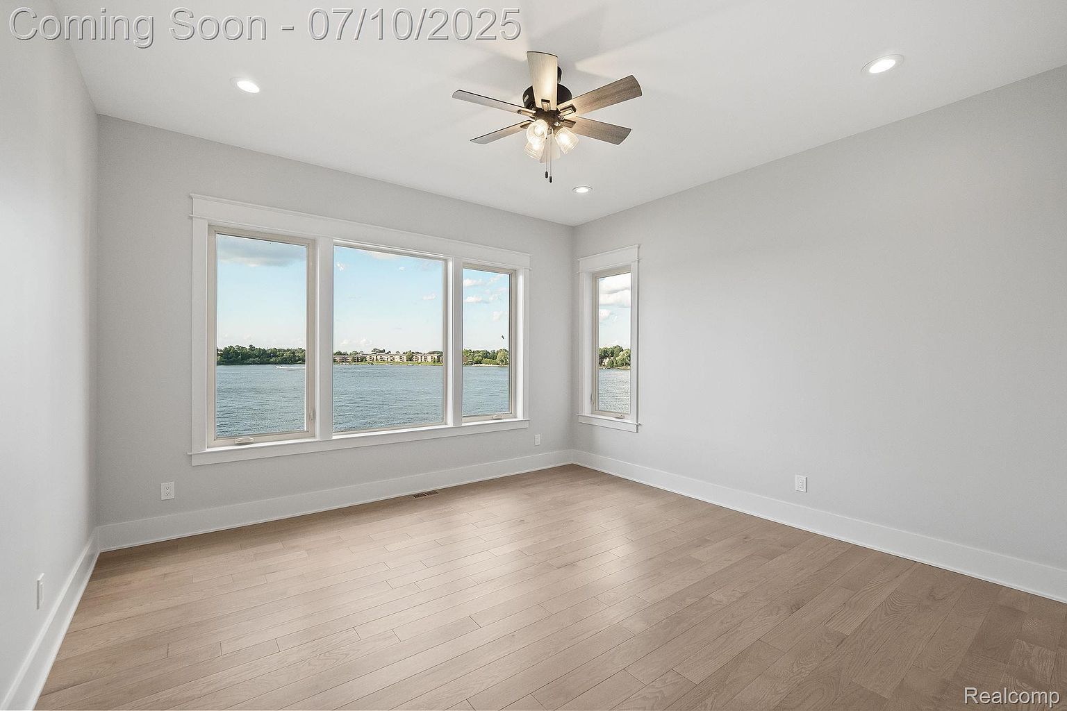 This is an interior shot of a bedroom featuring light hardwood floors and white walls. The room has large windows offering a view of a lake or river, and a ceiling fan provides air circulation. The space is bright and airy, creating a serene and inviting atmosphere.