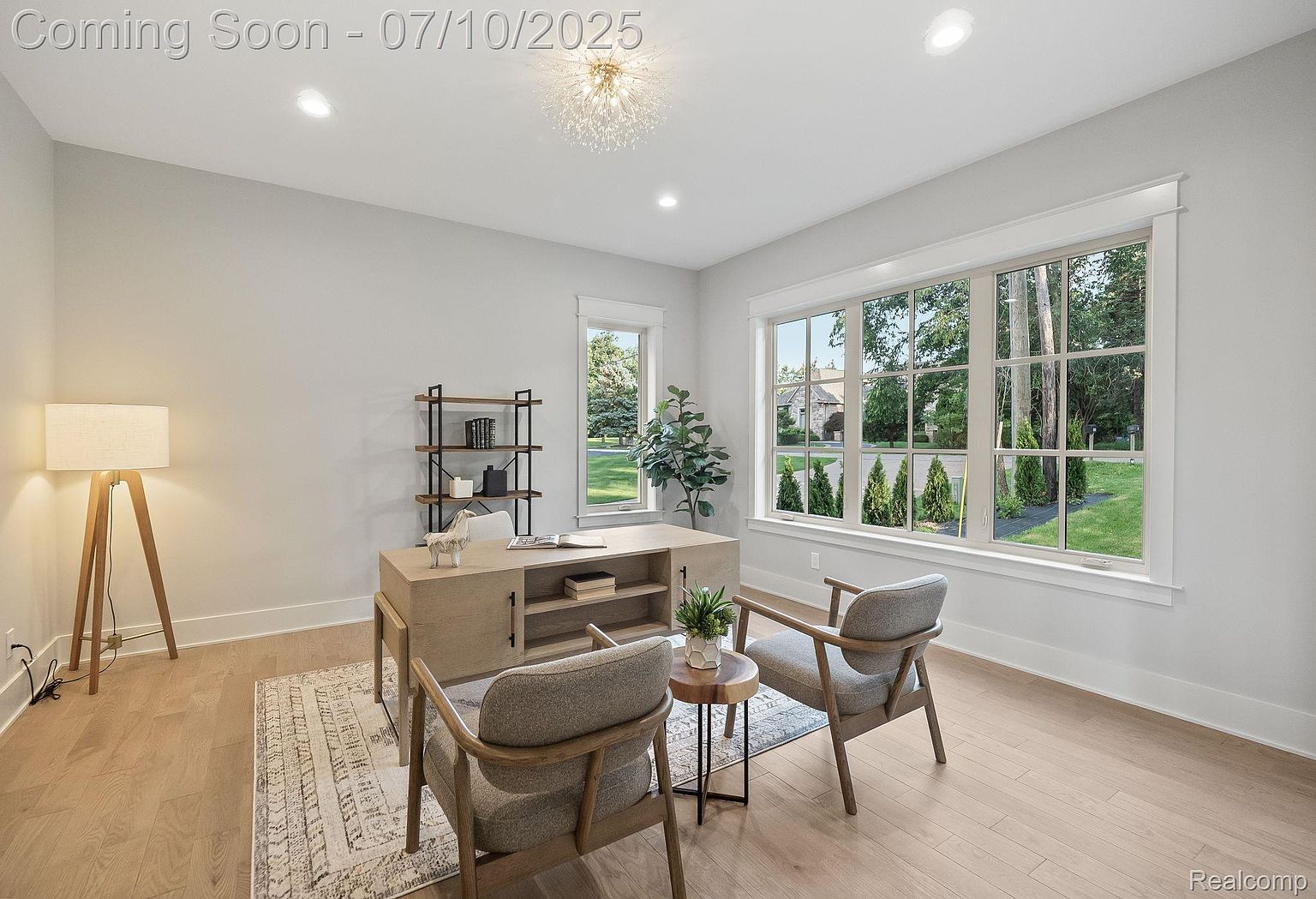 This is an interior shot of a modern office or study. The room features a light wood floor, a large window providing natural light, a desk with shelving, and two chairs with a small table between them. The overall impression is clean, bright, and inviting, perfect for a home office space.