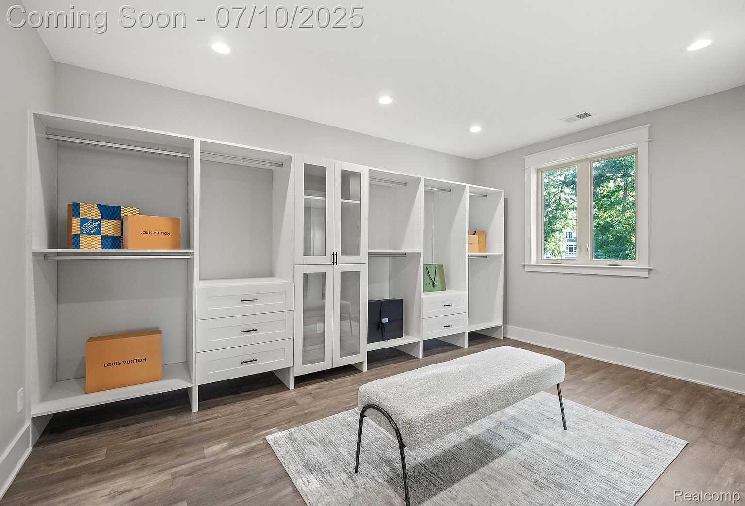 This is a well-organized walk-in closet featuring custom white shelving and cabinetry. The closet includes drawers, hanging rods, and glass-front cabinets, providing ample storage space. A plush bench sits on a light gray rug in the center of the room, adding a touch of luxury.