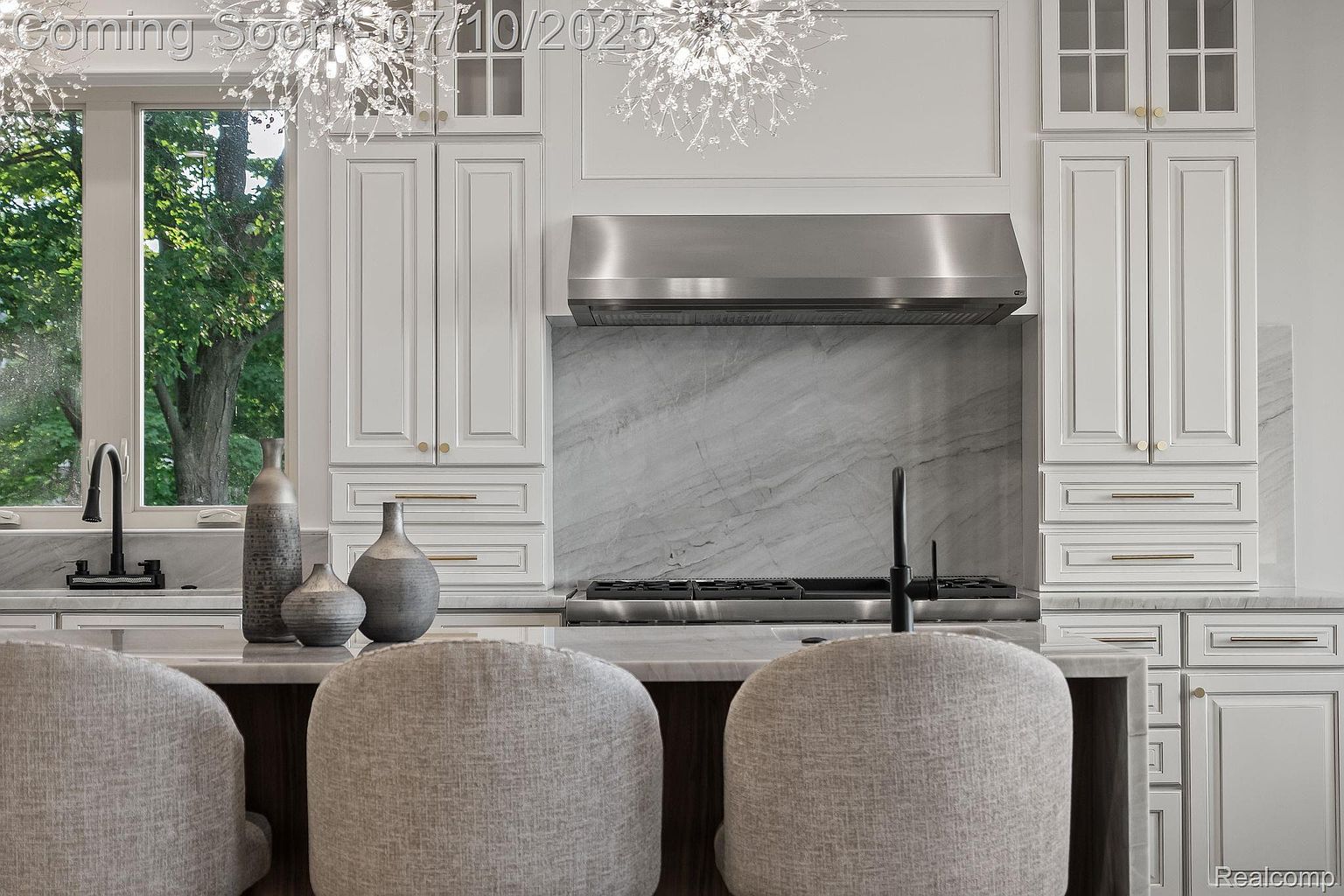 This is a bright and modern kitchen featuring white cabinetry with gold hardware, a stainless steel range hood, and a marble backsplash. A large island with seating provides a focal point, complemented by stylish light fixtures. The kitchen exudes a clean and sophisticated aesthetic, perfect for a contemporary home.