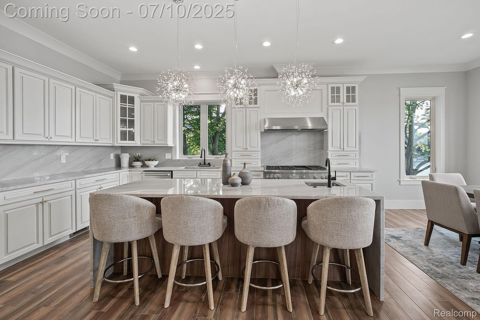 This is a bright and modern kitchen featuring white cabinetry, marble countertops, and stainless steel appliances. A large island with seating provides a central gathering space, illuminated by three decorative pendant lights. The hardwood flooring adds warmth to the space, creating an inviting atmosphere.