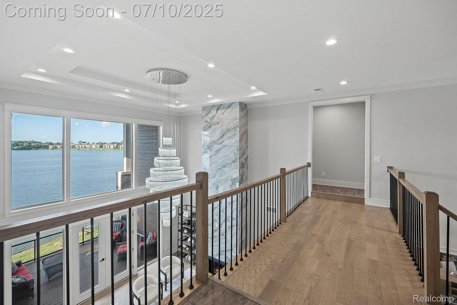 This interior shot showcases a modern hallway with hardwood flooring and a stylish staircase featuring dark metal railings and light wood posts. A large chandelier hangs in the center, adding a touch of elegance, while a window offers a view of the water. The space is well-lit and appears spacious, creating a luxurious and inviting atmosphere.