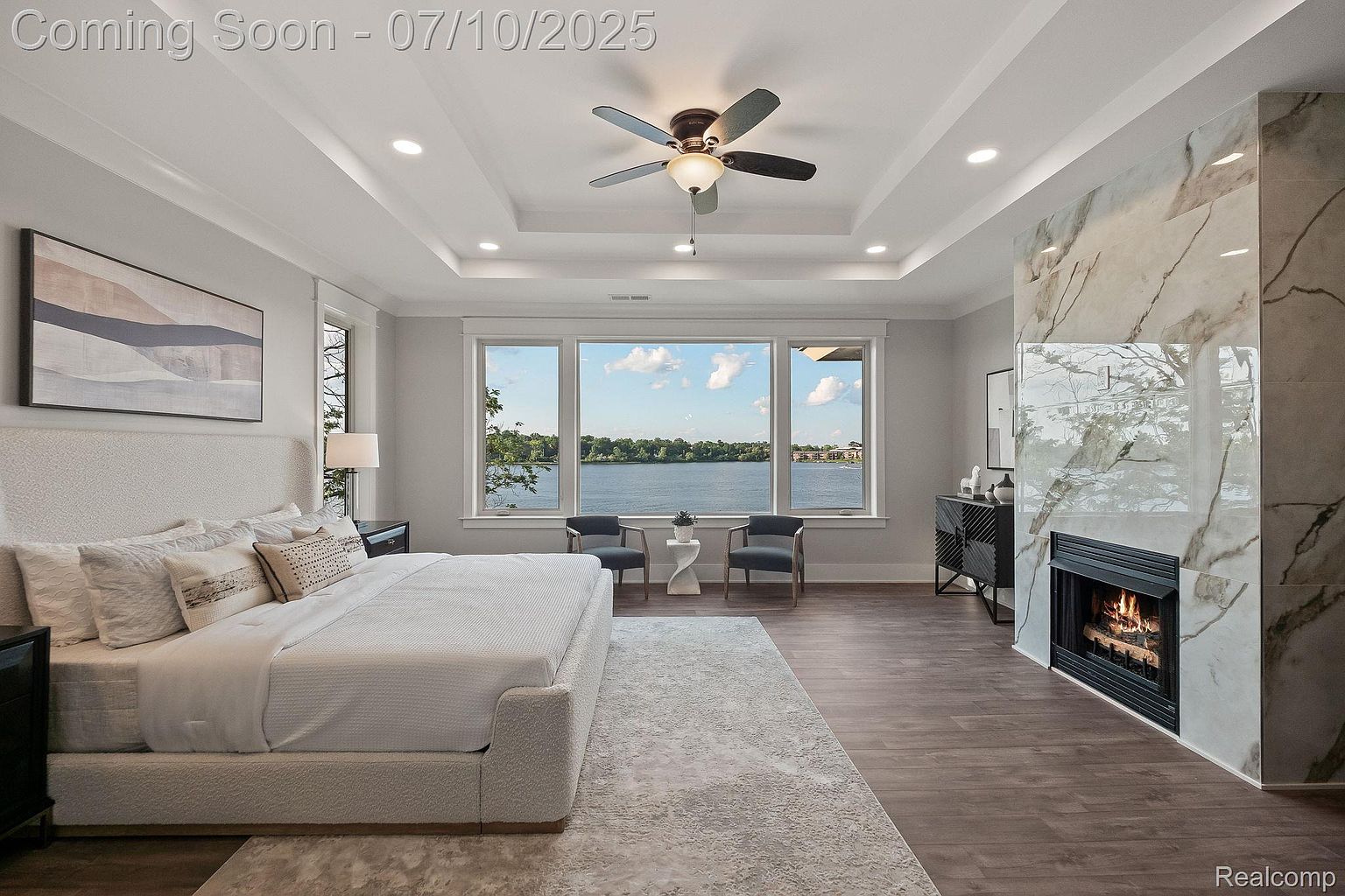 This is a luxurious primary bedroom featuring a large bed with a textured headboard and plush bedding, a modern fireplace with marble surround, and expansive windows offering a serene water view. The room is well-lit with recessed lighting and a ceiling fan, creating a bright and inviting atmosphere. The dark wood flooring and neutral color palette contribute to a sophisticated and relaxing ambiance.