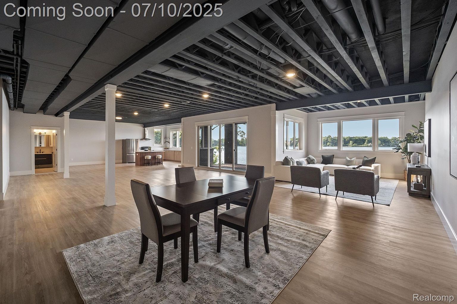 This is a spacious, open-concept living area featuring a dining space, a living room, and a glimpse into the kitchen. The room boasts modern design elements, including a dark exposed ceiling, light wood flooring, and large windows offering a view of the water. The space is well-lit and appears to be designed for comfortable living and entertaining.