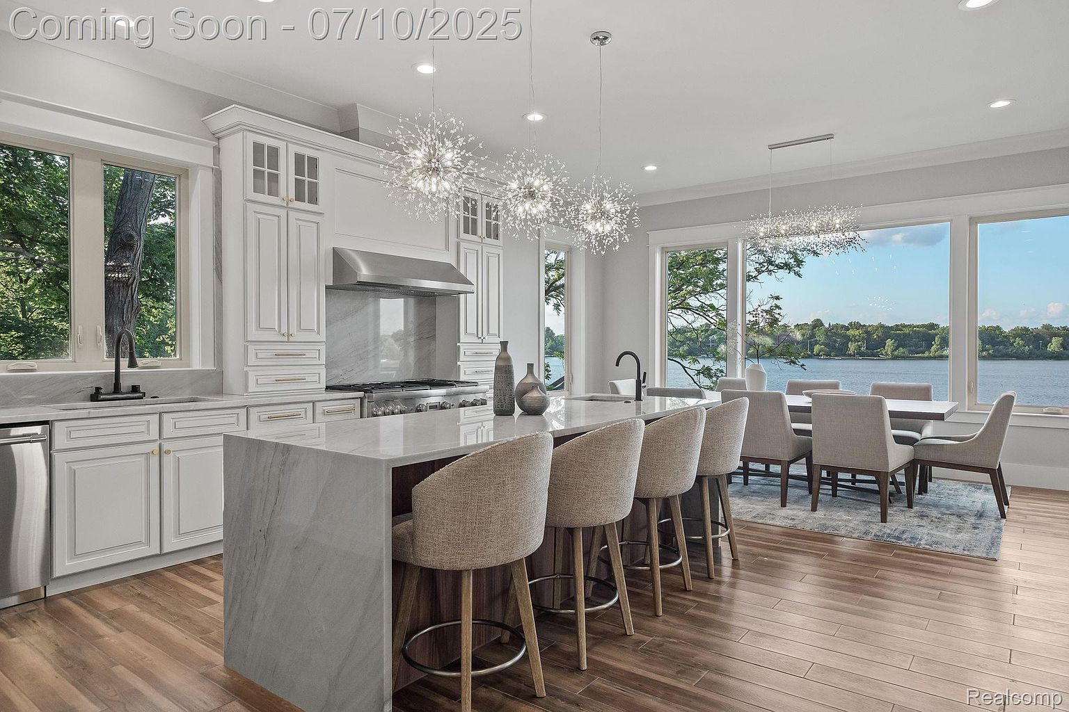This is a bright and modern kitchen featuring white cabinetry, a large marble island with seating, and stainless steel appliances. The kitchen is well-lit with unique pendant lighting and natural light from the large windows overlooking a lake. A dining area is visible adjacent to the kitchen, creating an open and inviting space.