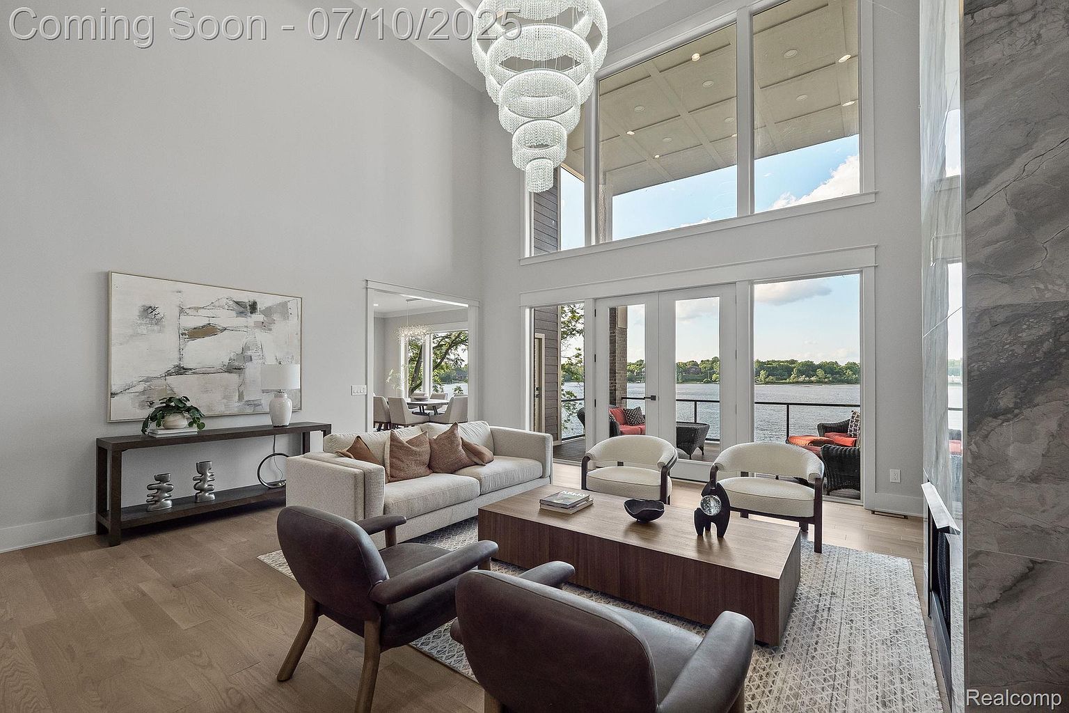 This is a spacious living room with high ceilings and large windows offering a view of the water. The room features a modern chandelier, a neutral-toned sofa with decorative pillows, and two leather armchairs facing a wooden coffee table. An abstract painting hangs above a console table, adding to the room's contemporary aesthetic.