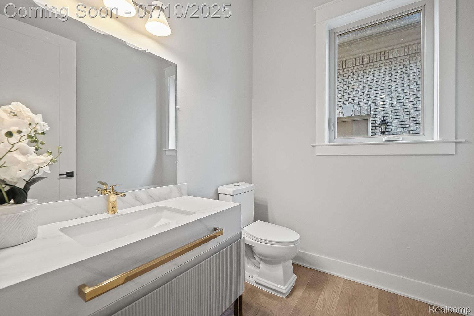 This is a well-lit bathroom featuring a modern vanity with a white countertop and gold hardware. A sleek, gold faucet complements the sink, and a toilet is positioned next to a window that provides natural light. The overall aesthetic is clean, bright, and contemporary, creating a welcoming and stylish space.