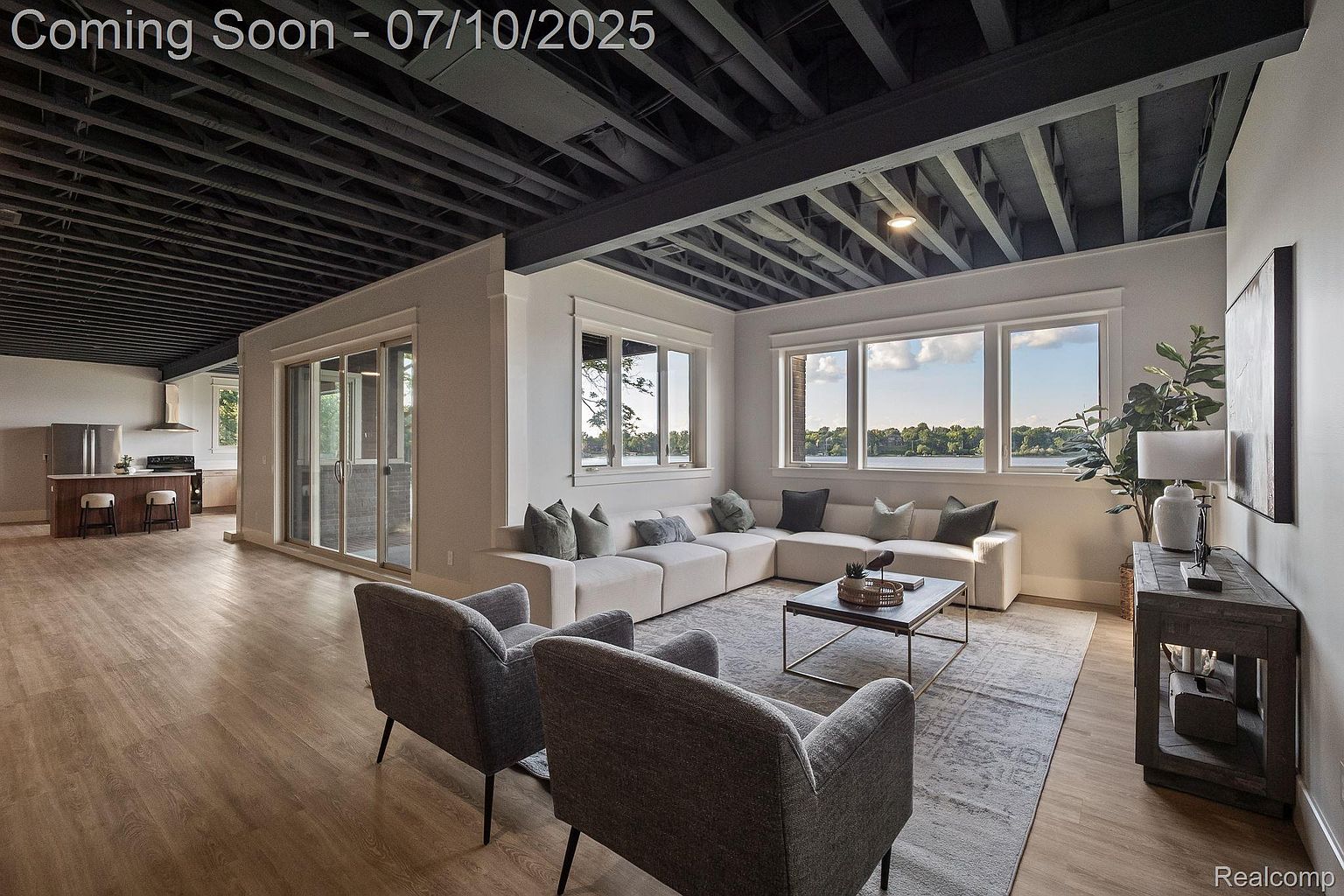 This is a spacious living room featuring an open floor plan with a modern design. The room includes a large sectional sofa, two armchairs, a coffee table, and a console table with a lamp. Large windows provide ample natural light and views of the outdoors, while the exposed ceiling beams add an industrial touch.