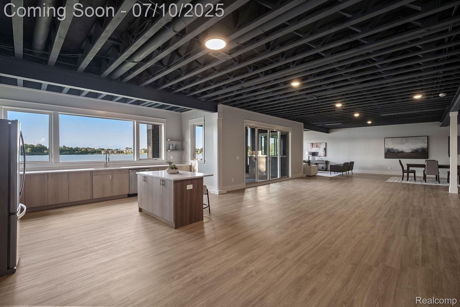 This interior shot showcases a spacious, modern kitchen and living area. The kitchen features light wood cabinetry, a central island, and large windows overlooking a waterfront view. The open floor plan extends into a living area with contemporary furniture and artwork, creating a seamless and inviting space.