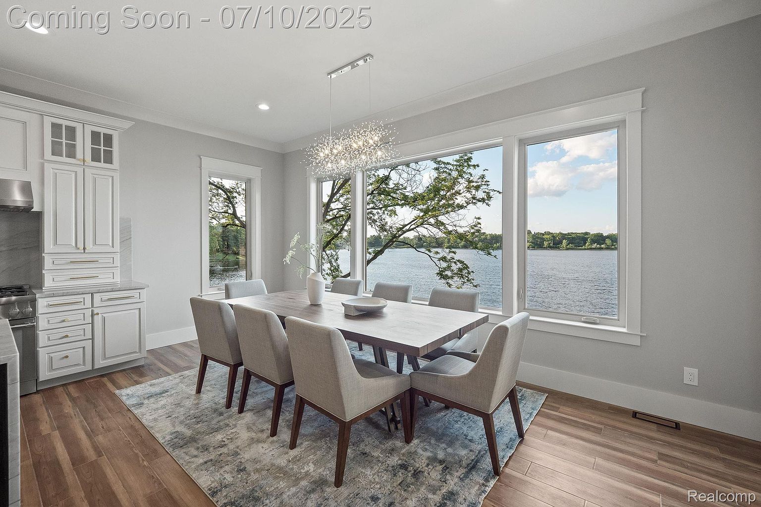 This is an interior shot of a dining room featuring a large wooden table with six upholstered chairs. A modern chandelier hangs above the table, and a large window offers a scenic view of a lake. The room is decorated in neutral tones, creating a bright and airy atmosphere.