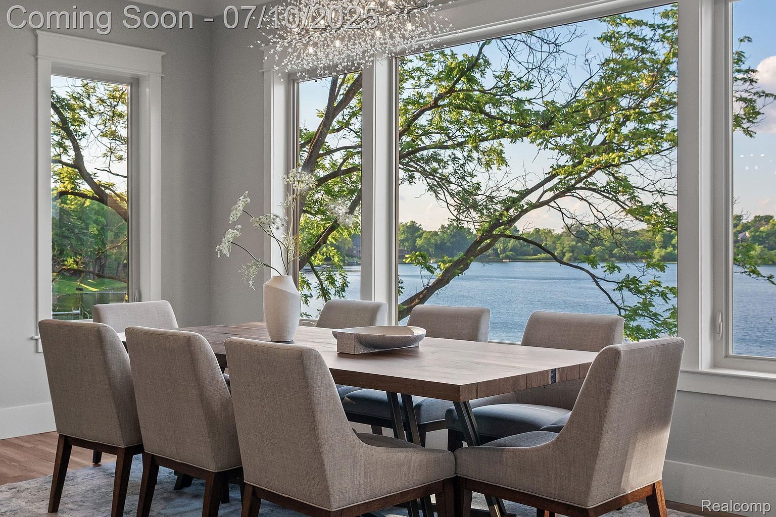 This is an interior shot of a dining room featuring a large wooden table surrounded by upholstered chairs. A modern chandelier hangs above the table, and large windows offer a scenic view of a lake and trees. The room is well-lit and has a neutral color palette, creating a serene and elegant atmosphere.