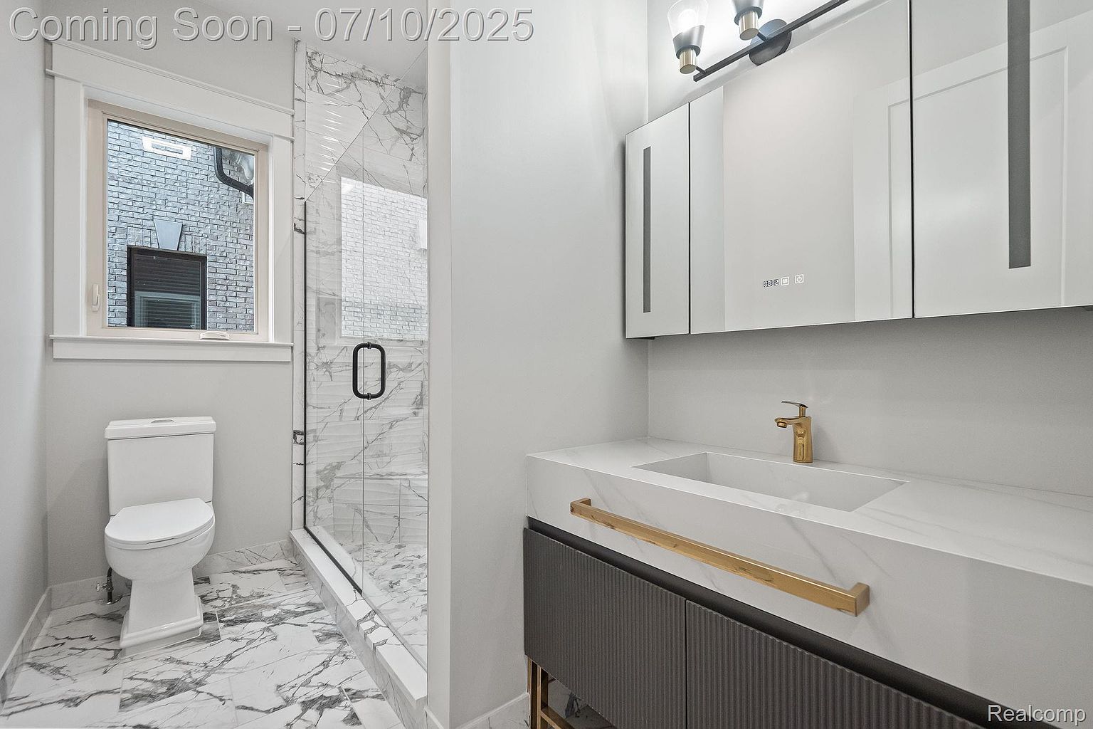 This is a modern bathroom featuring a toilet, a glass-enclosed shower with marble-like tiling, and a floating vanity with a marble countertop and gold hardware. The color palette is primarily white and gray, creating a clean and sophisticated look. The image is taken from a wide angle to showcase the entire space.
