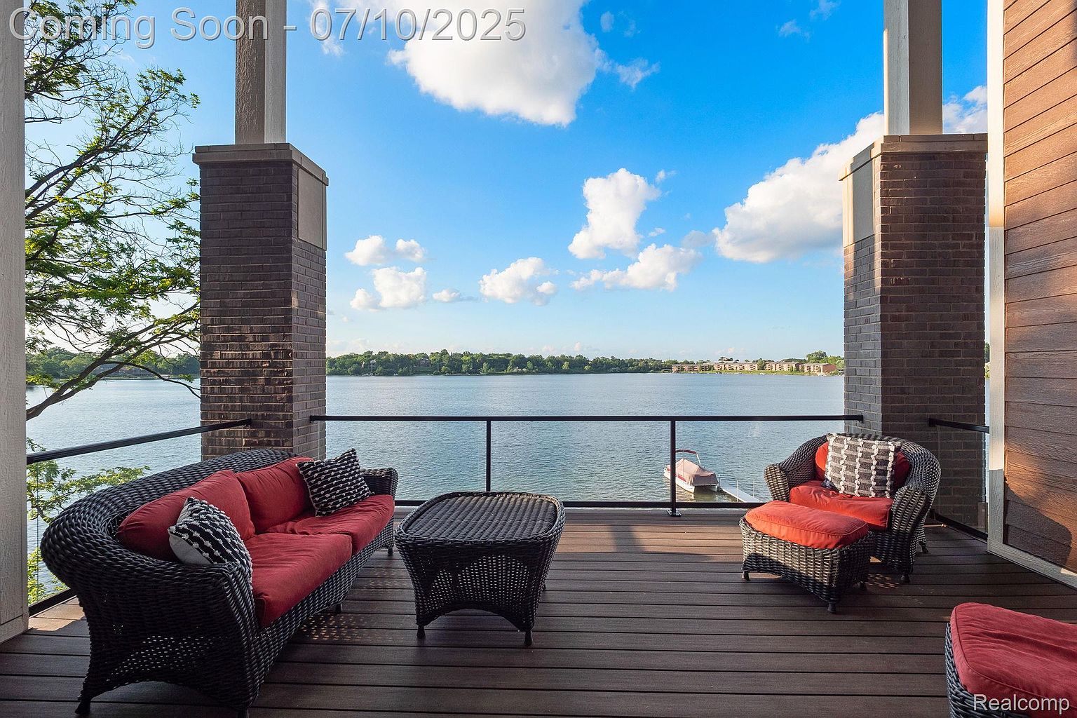 This image showcases a beautifully furnished outdoor patio or balcony with a stunning waterfront view. The space features comfortable wicker furniture with red cushions, including a sofa, armchair, and coffee table, all arranged on a wooden deck. The view overlooks a serene lake with distant buildings and a clear blue sky with scattered clouds, creating a relaxing and luxurious outdoor living space.