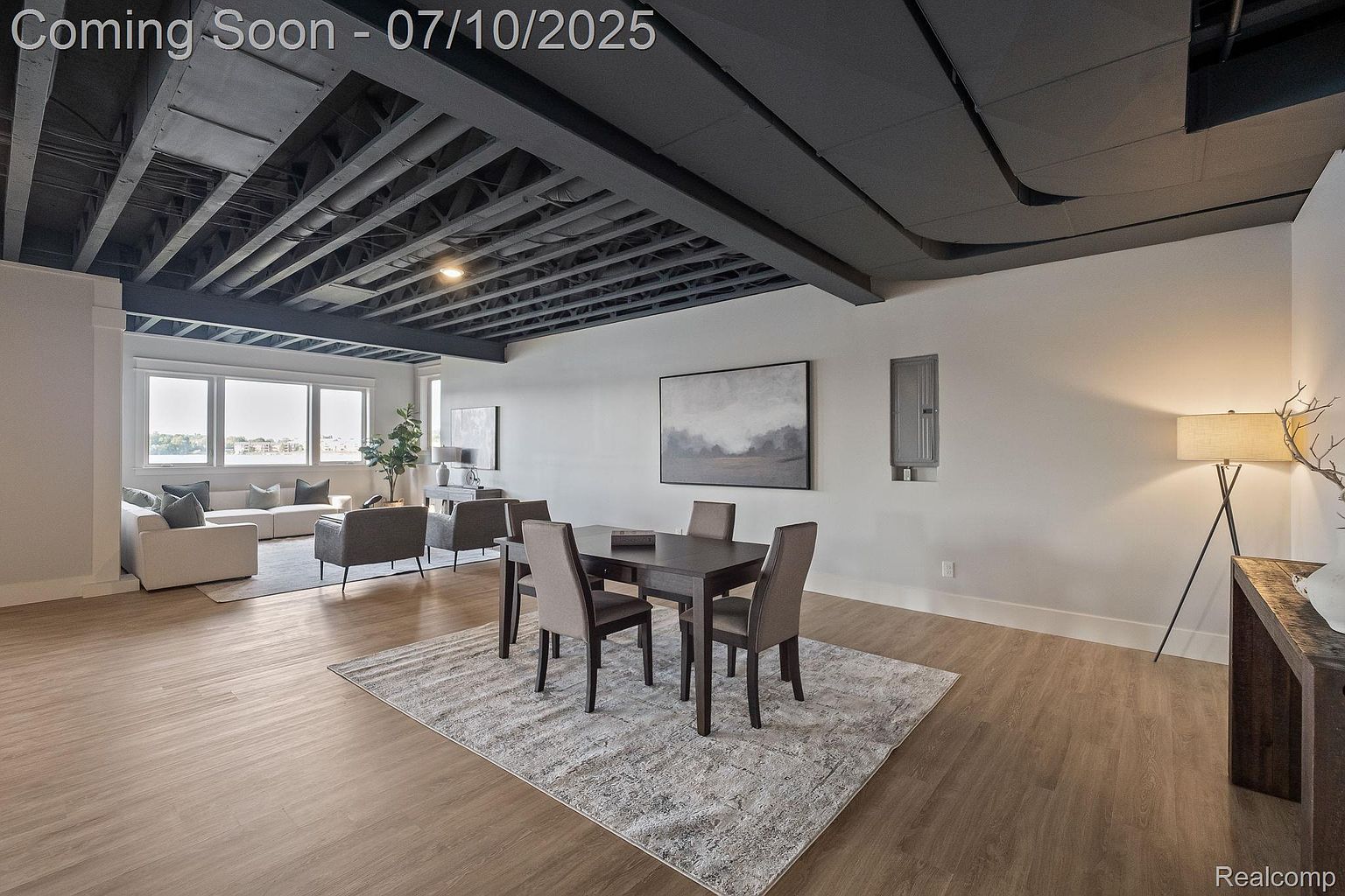 This interior shot showcases a modern living and dining area with an exposed ceiling painted in a dark color. The space features a seating area with a sofa and chairs near large windows, a dining table with chairs on a patterned rug, and light wood-look flooring throughout. The overall impression is a stylish and open living space.
