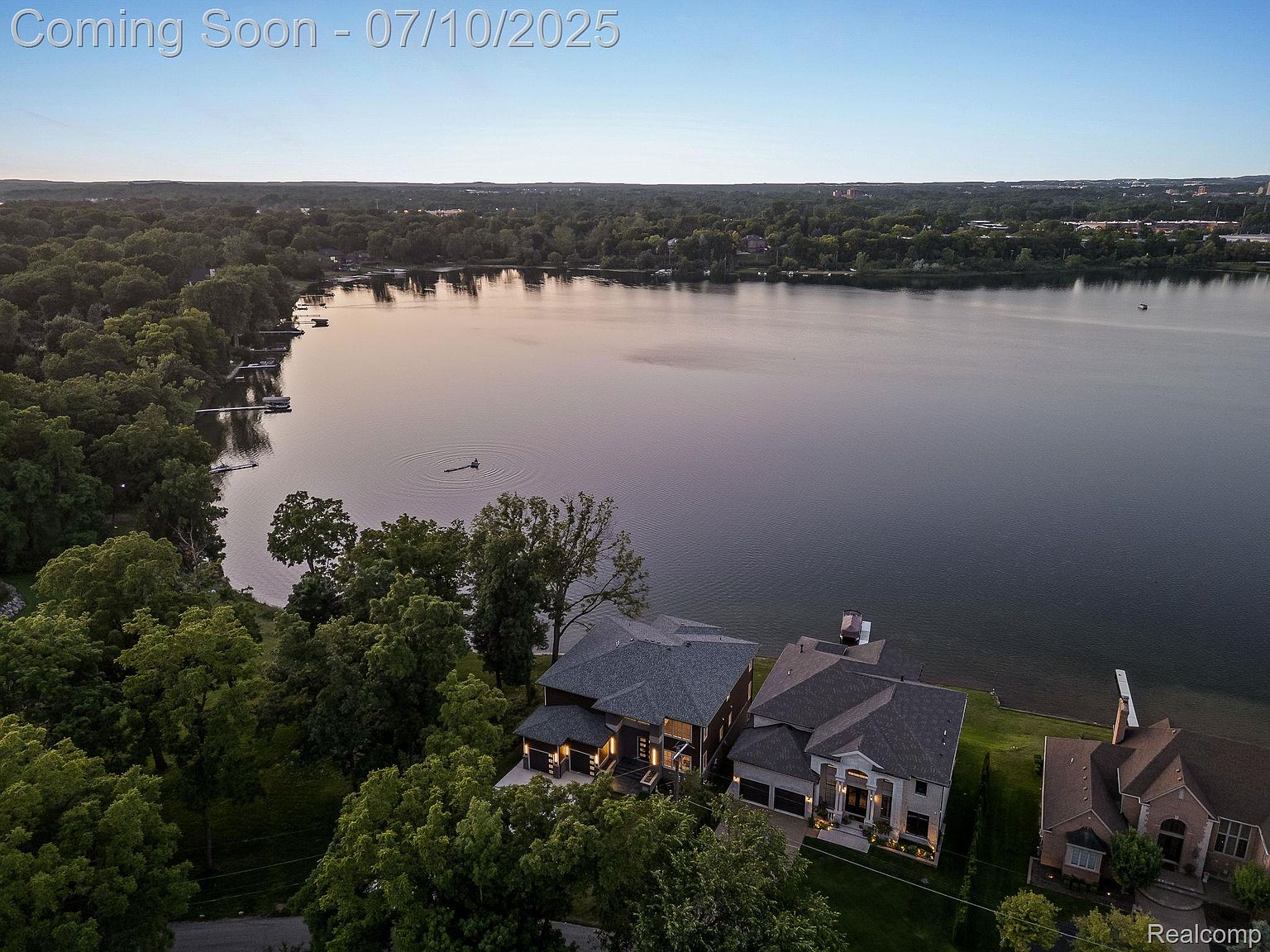 This aerial view showcases a stunning lakeside property featuring multiple houses with well-maintained lawns extending to the water's edge. The homes exhibit modern architectural styles with gray roofs and neutral-toned exteriors. Lush greenery surrounds the properties, creating a serene and private atmosphere, while the expansive lake provides a picturesque backdrop.