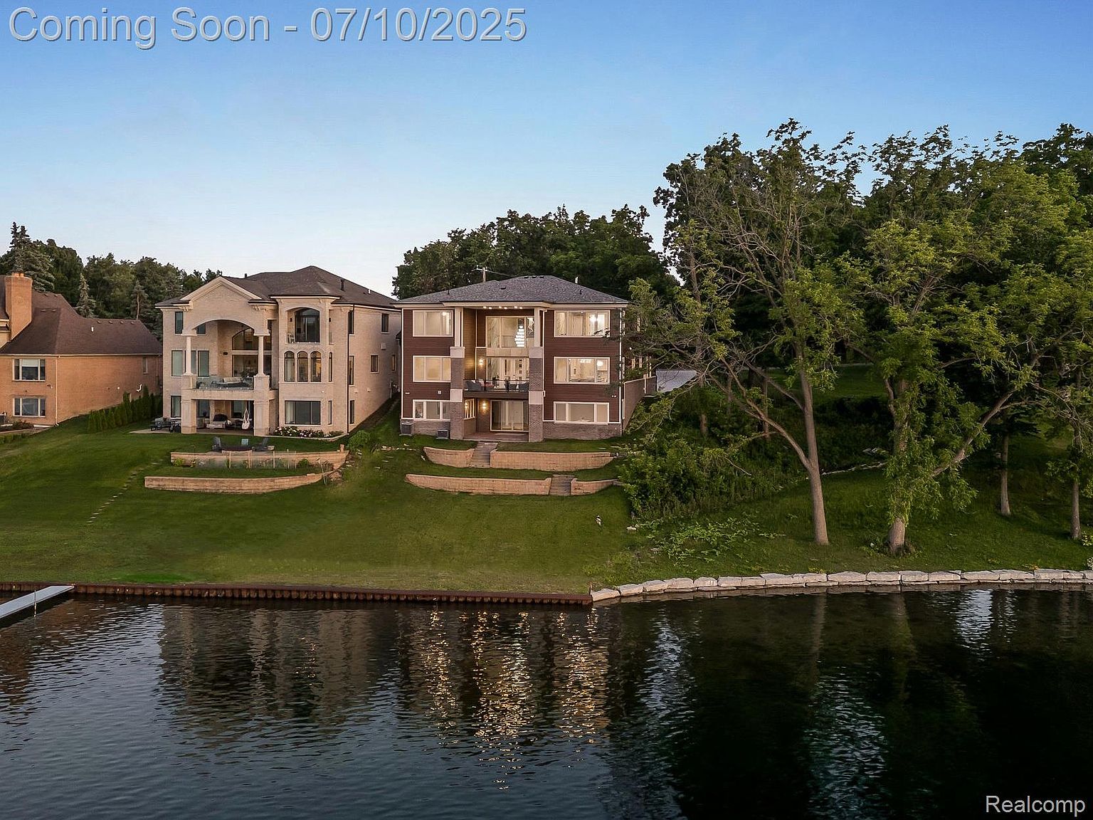 This aerial view showcases two luxurious waterfront homes with manicured lawns and stone retaining walls along the water's edge. The homes feature multi-story designs with large windows and balconies, offering stunning views of the water. Lush trees surround the properties, adding to the sense of privacy and exclusivity.