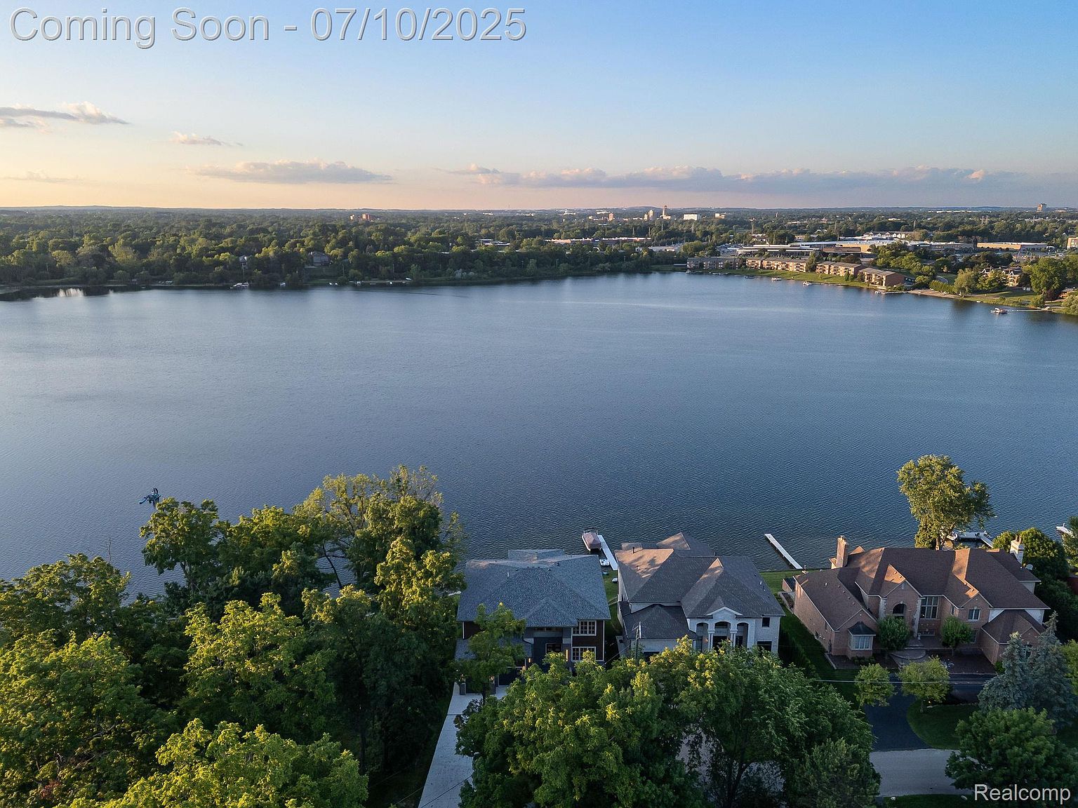 This aerial view showcases a stunning lakeside property with multiple houses nestled among lush greenery. The expansive lake dominates the scene, reflecting the sky and creating a serene atmosphere. The houses feature varied architectural styles, each with private docks extending into the water, suggesting a luxurious and tranquil living environment.