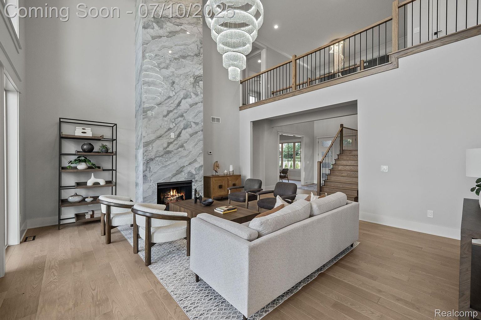This is an interior shot of a modern living room featuring a high ceiling, a marble fireplace, and a large chandelier. The room is furnished with a light-colored sofa, two armchairs, and a wooden coffee table, all arranged on a patterned rug. A staircase is visible in the background, leading to a second-floor balcony overlooking the living room, creating an open and airy atmosphere.
