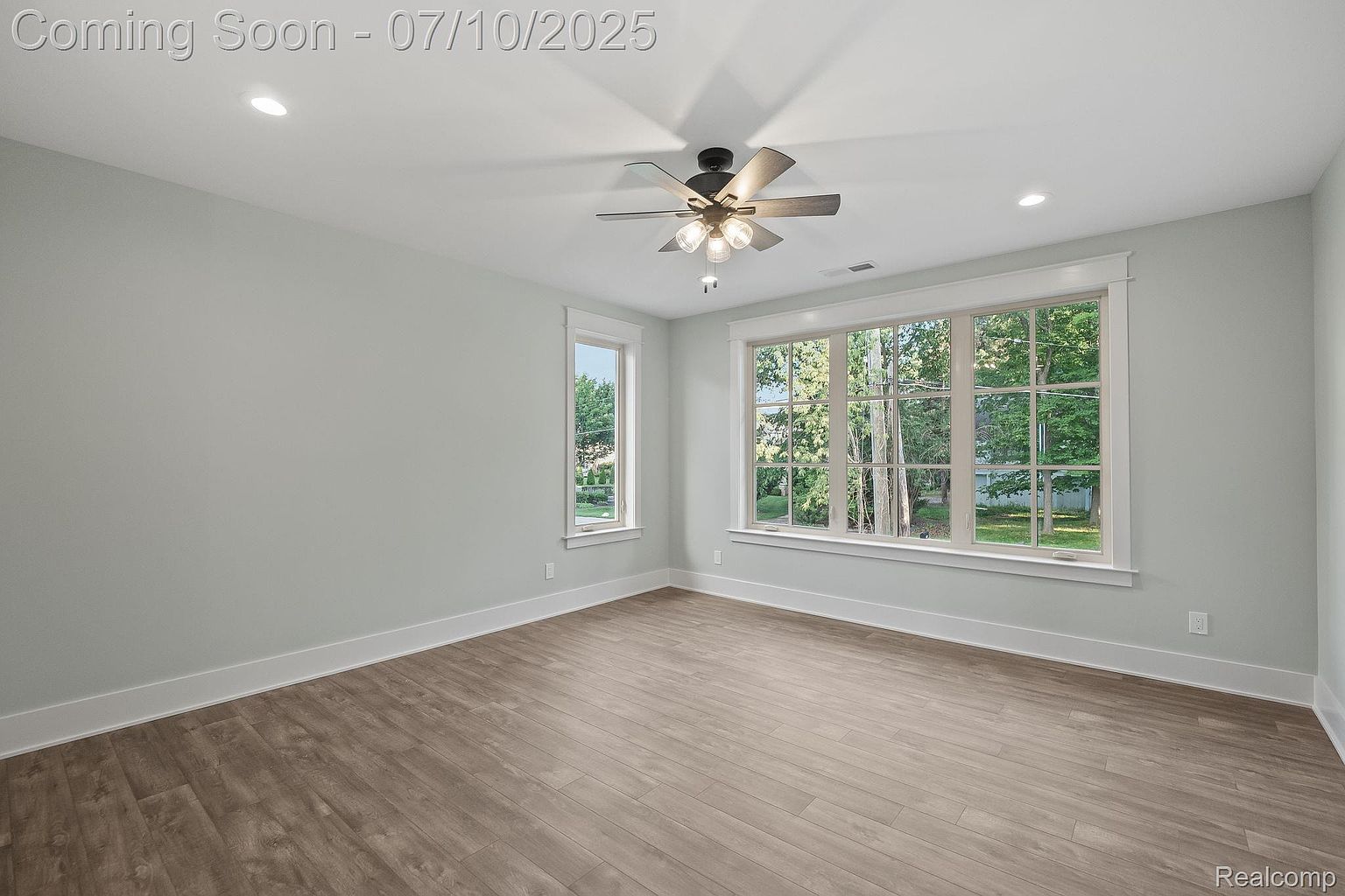 This is an interior shot of a bedroom featuring light wood flooring and light blue walls. A large window provides ample natural light, complemented by a ceiling fan with lights. The room is spacious and appears to be newly constructed or renovated, giving a clean and modern impression.