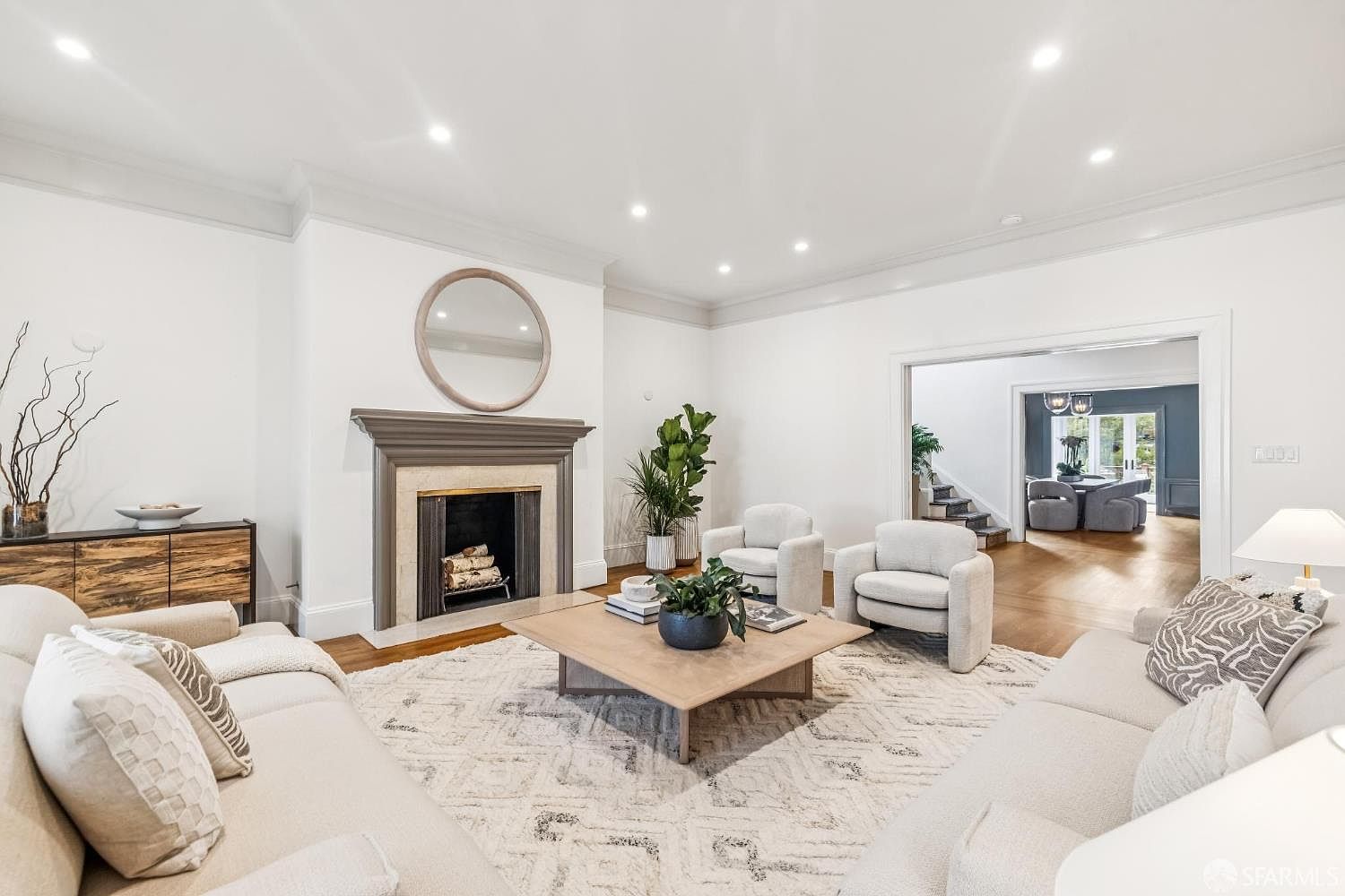 This is a bright and spacious living room featuring a neutral color palette, a fireplace with a round mirror above, and comfortable seating arrangements. The room is well-lit with recessed lighting and natural light, and a large area rug anchors the space. The open doorway leads to another room, creating a sense of flow and connectivity within the home.