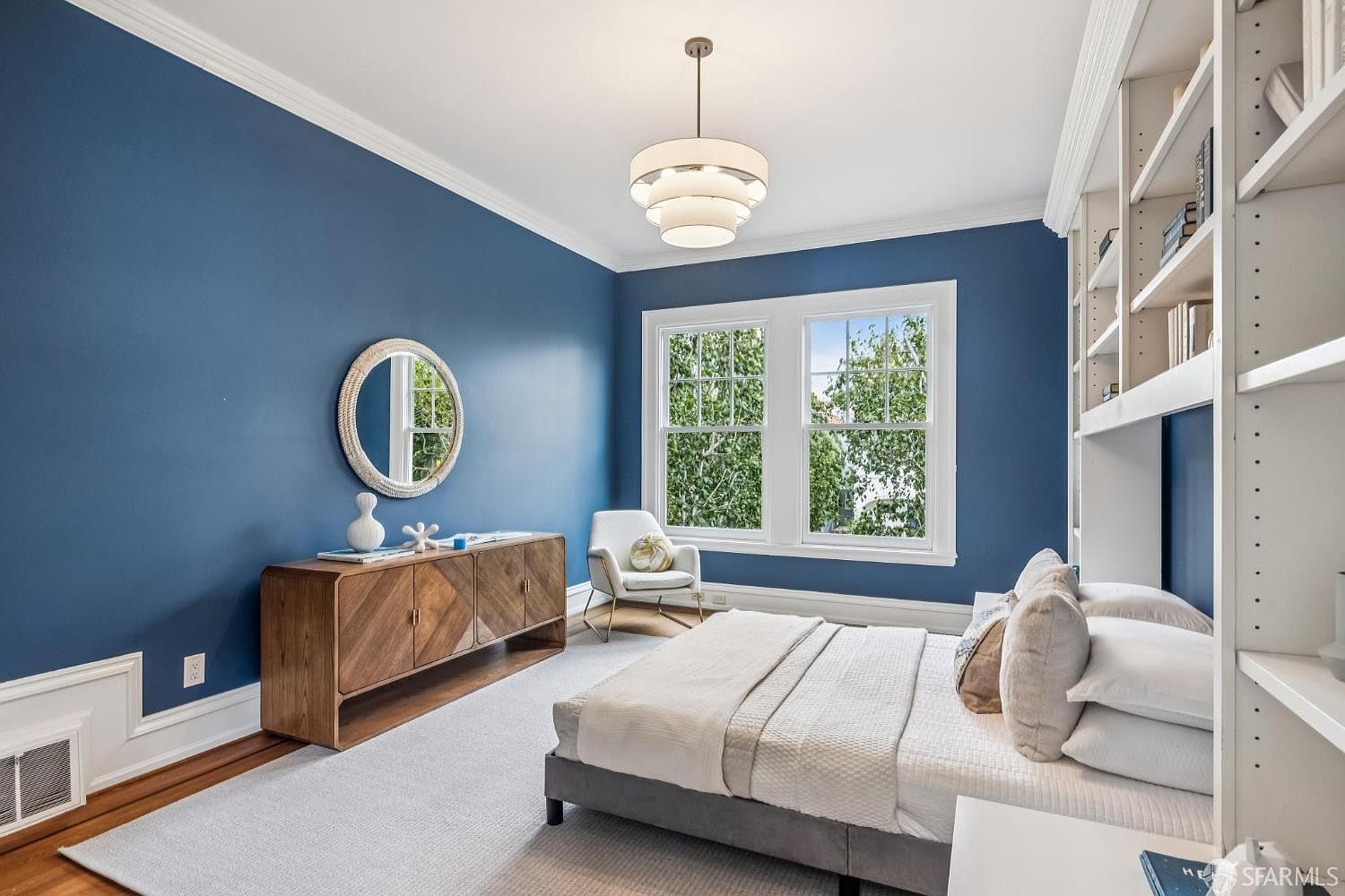 This is a beautifully decorated primary bedroom featuring a striking blue accent wall, complemented by white trim and a large window offering natural light. The room includes a modern bed with neutral bedding, a stylish wooden dresser with decorative items, and a round mirror. A contemporary light fixture hangs from the ceiling, adding to the room's sophisticated ambiance.