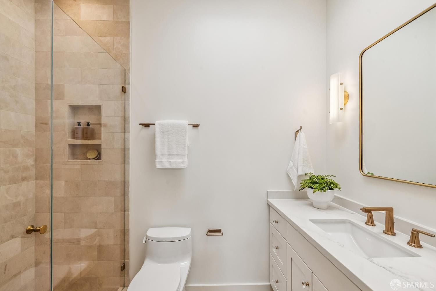 This is a well-lit guest bathroom featuring a walk-in shower with beige tiling and a glass door. The bathroom includes a white toilet, a vanity with a white countertop and white cabinets, and a gold-framed mirror. A small green plant adds a touch of freshness to the space.