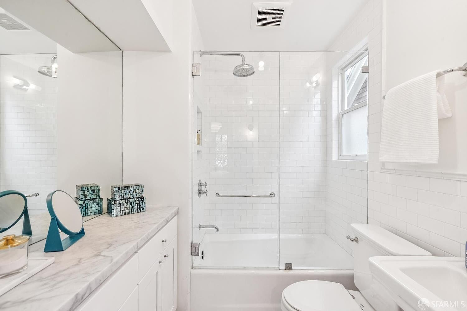 This is a bright and clean bathroom featuring white subway tile, a glass-enclosed shower/tub combination, and a white vanity with a marble countertop. The room is well-lit, with natural light coming through a window above the toilet. The overall impression is fresh and modern.