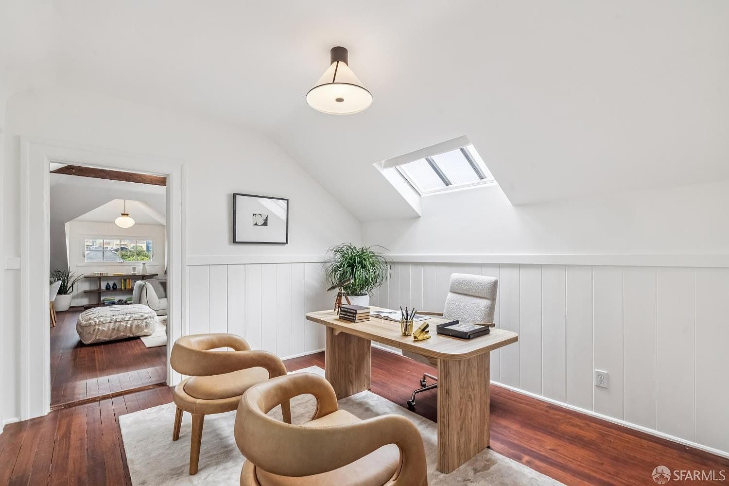 This is an interior shot of a bright and airy home office. The room features a wooden desk with a white chair, two tan leather chairs, and a skylight that allows natural light to flood the space. The walls are white with wainscoting, and the hardwood floors add warmth to the room.