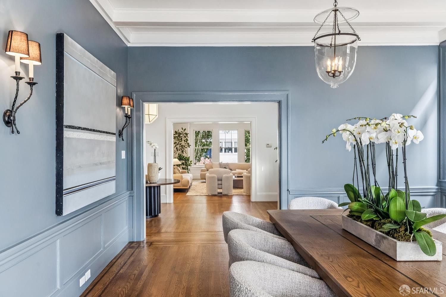 This is an interior shot of a dining room, showcasing a blend of classic and contemporary design elements. The room features blue walls with wainscoting, a large abstract painting, and sconce lighting. A wooden dining table with upholstered chairs is the focal point, adorned with a large floral arrangement. The view through the doorway reveals a glimpse into a bright living room, creating a sense of spaciousness and flow.