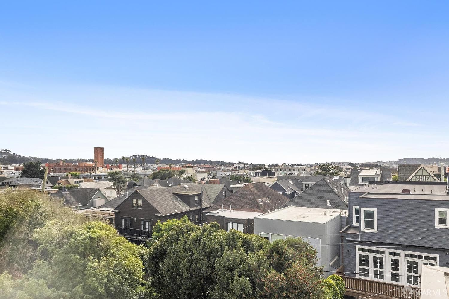 This aerial view showcases a residential neighborhood with a mix of architectural styles, featuring houses with varying rooflines and colors. Lush greenery surrounds the homes, adding to the neighborhood's charm. In the distance, a brick tower and the city skyline are visible under a clear blue sky, providing a sense of location and scale.