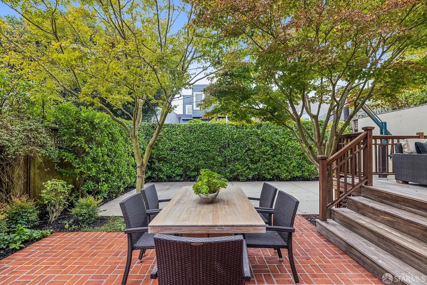 This is a well-maintained backyard featuring a brick patio with an outdoor dining set, including a wooden table and wicker chairs. Lush green hedges and trees provide privacy and create a serene atmosphere. A wooden deck with stairs is visible on the right, suggesting an easy transition between indoor and outdoor living spaces.