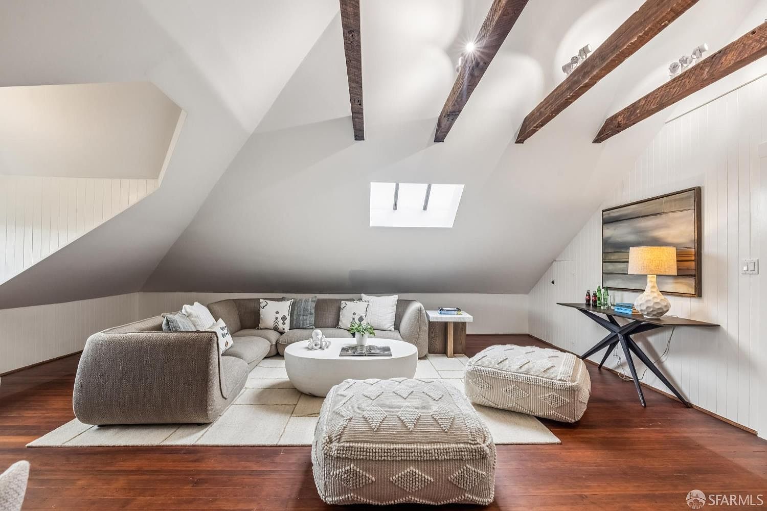 This is an interior shot of a living room in an attic space. The room features a sectional sofa, a round coffee table, and two poufs on a light-colored rug. The ceiling has exposed wooden beams and a skylight, adding character to the space. A console table with a lamp and artwork is placed against a white paneled wall.