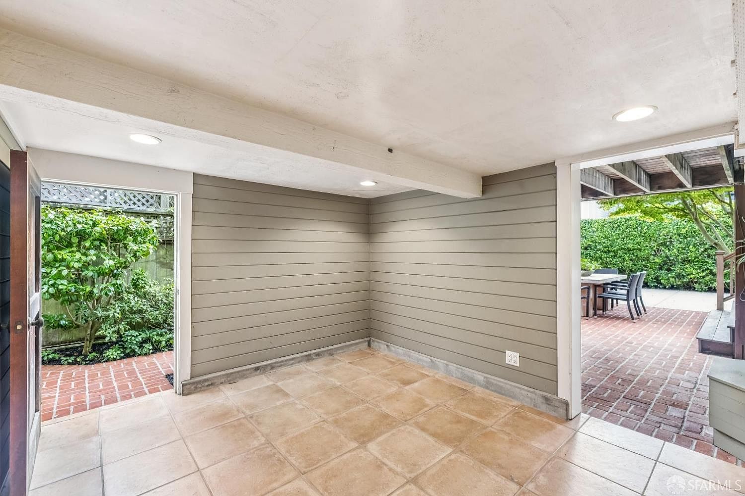This sunroom features a tiled floor and horizontally paneled walls, creating a bright and airy space. An open doorway leads to a brick patio and garden area, while another doorway opens to a lush green space. The room's design seamlessly blends indoor and outdoor living.